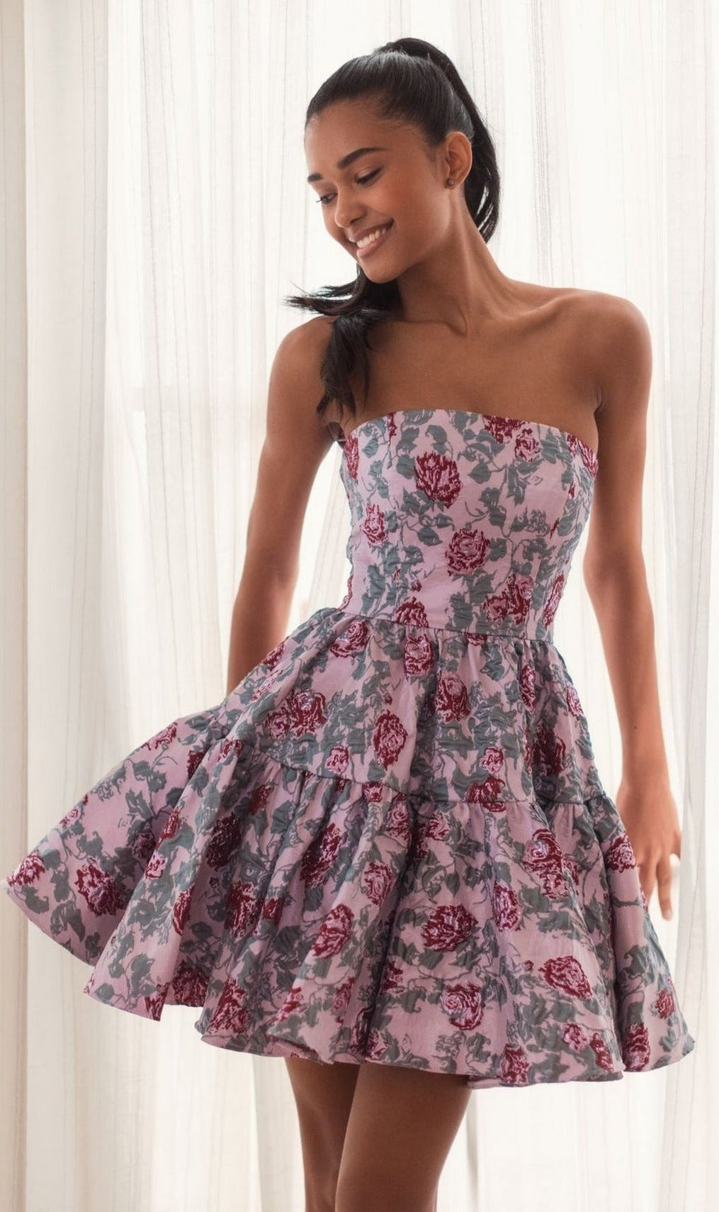 A woman smiles and looks down while wearing the Short Pink Floral Dress 68624 by Mac Duggal, featuring a strapless design with pink, grey, and red floral patterns, standing in front of sheer curtains.