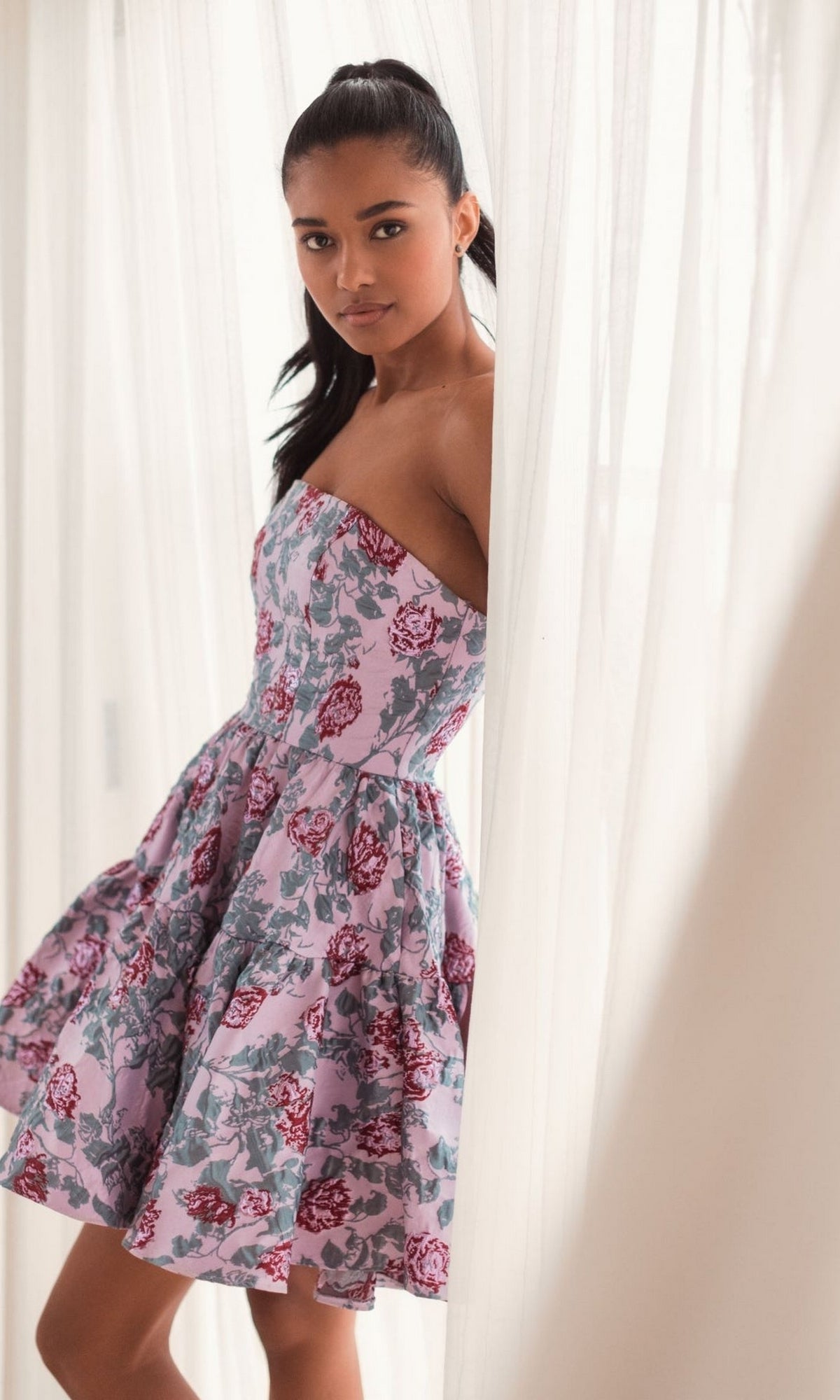 A woman in the Mac Duggal Short Pink Floral Dress 68624 with a floral print stands by white curtains, looking at the camera.