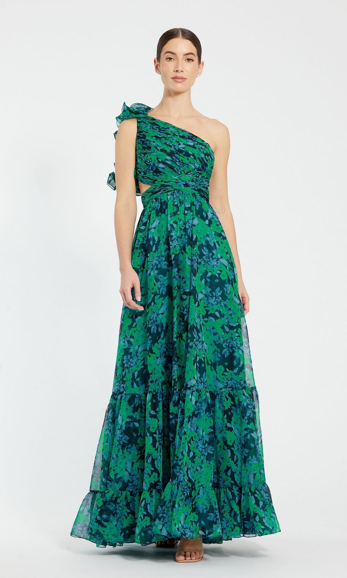 A woman stands against a white background wearing the Mac Duggal 68650 One-Shoulder Print Formal Dress, a long, sleeveless gown in green and blue floral print with ruffled detail.