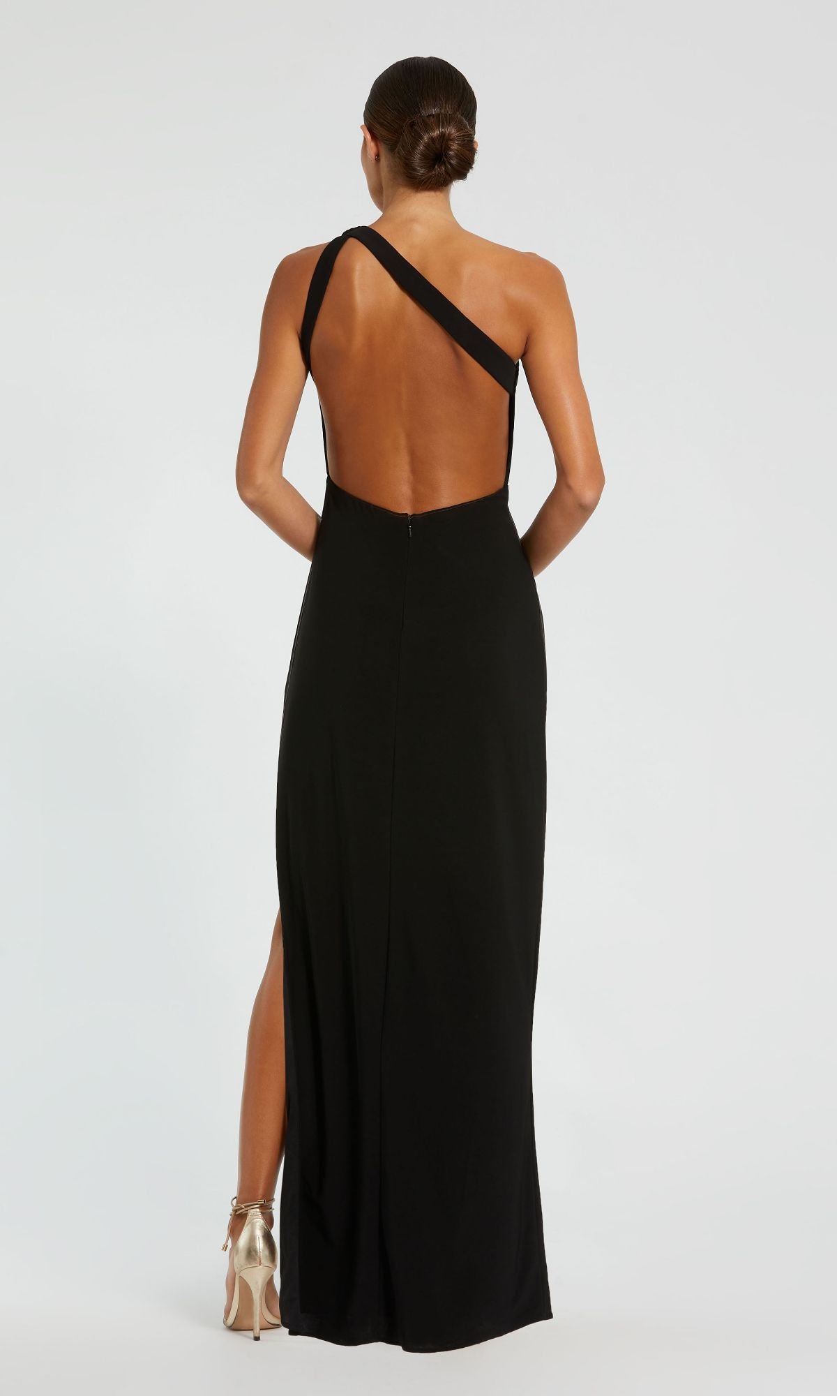 A person stands elegantly with their back to the camera, wearing the Mac Duggal 68690 One-Shoulder Black Prom Dress featuring an open back and high slit.