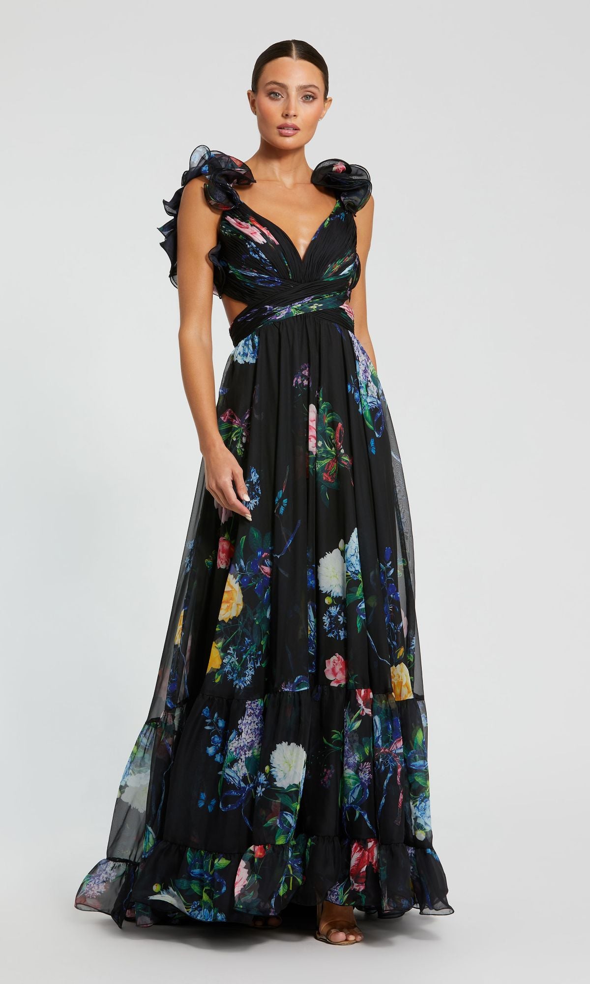 A woman wears the Mac Duggal 68746 Black Floral Formal Evening Gown, featuring ruffled shoulders, side cutouts, and a floor-length skirt, standing against a plain light background.