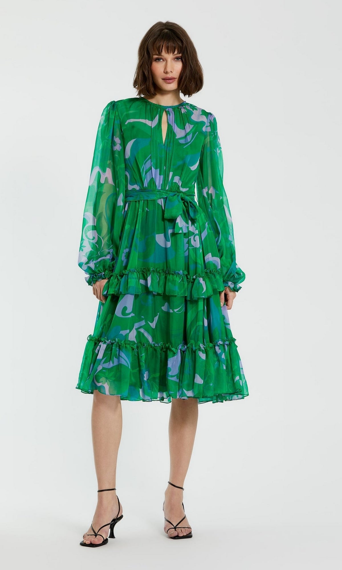 A woman models the Short Wedding Guest Dress 68760 by Mac Duggal, a green floral A-line dress with long sleeves, ruffled tiers, and a belted waist, styled with black strappy heels against a plain white background.