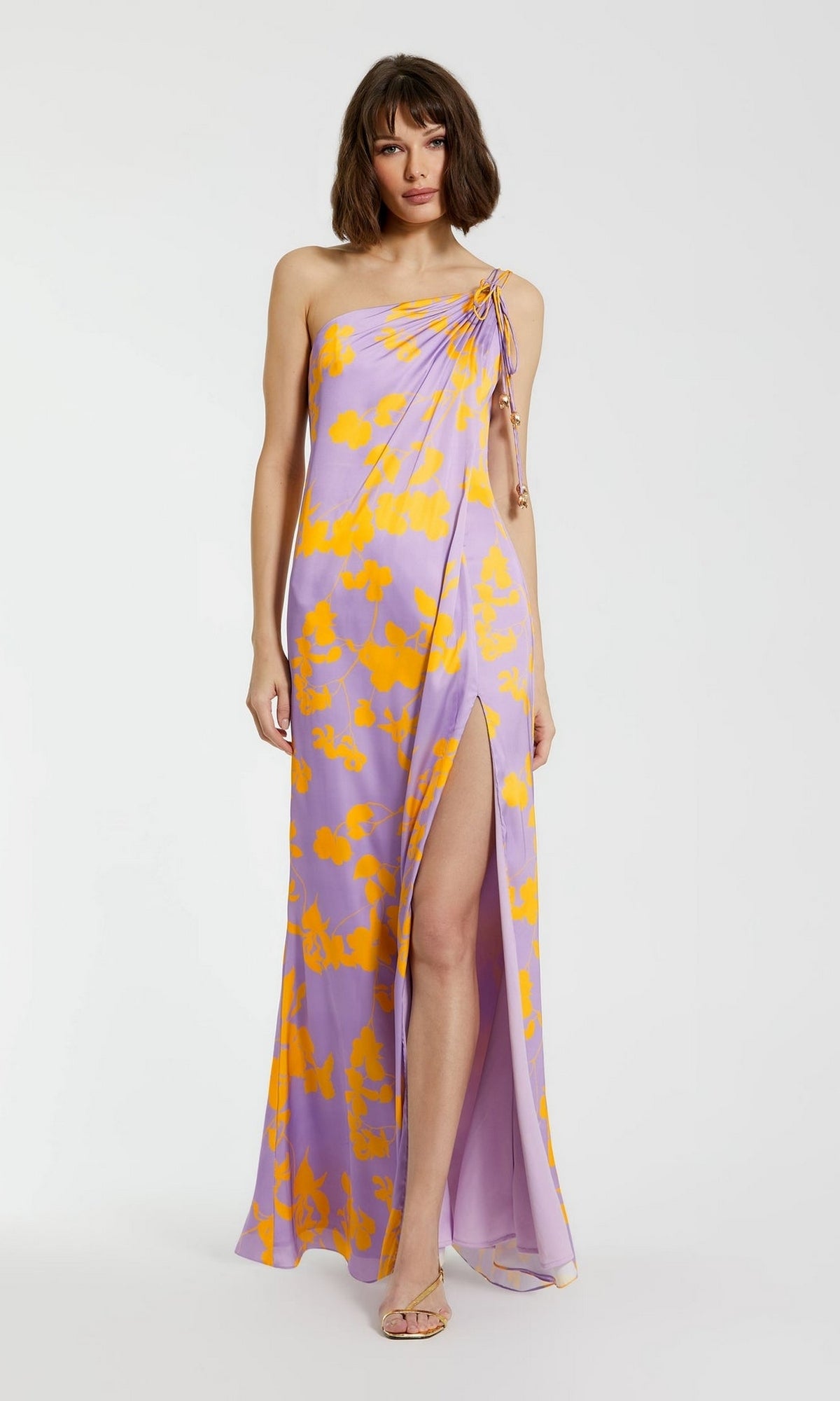 A woman wears the Mac Duggal Long Formal Dress 68771, a sleeveless one-shoulder lilac charmeuse gown with yellow abstract patterns and a high leg slit, standing against a plain background.