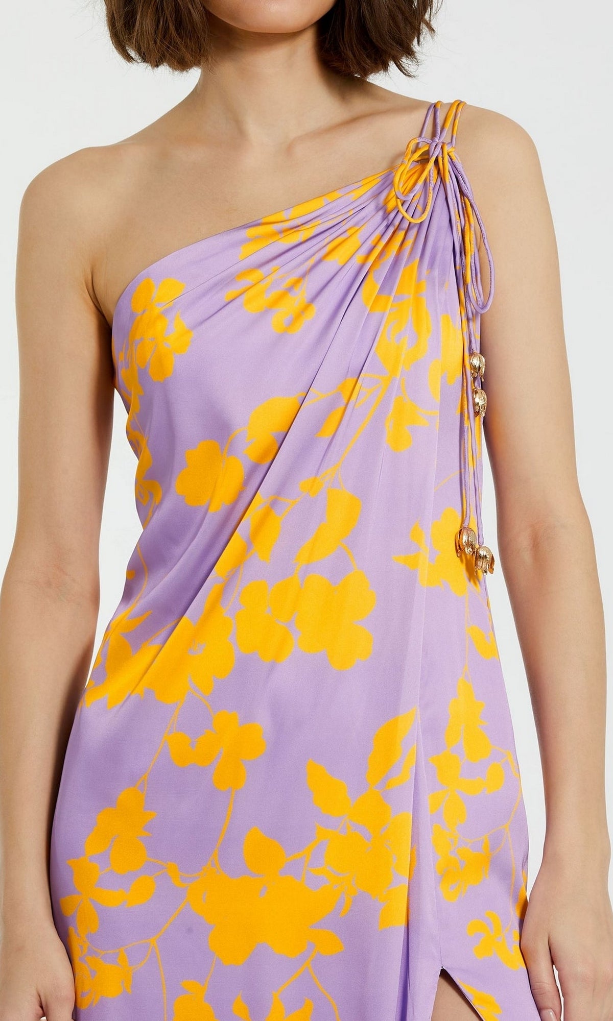 A woman wears the Long Formal Dress 68771 by Mac Duggal, a one-shoulder charmeuse gown with a purple base, bright yellow floral print, and gathered beaded detail at the shoulder.