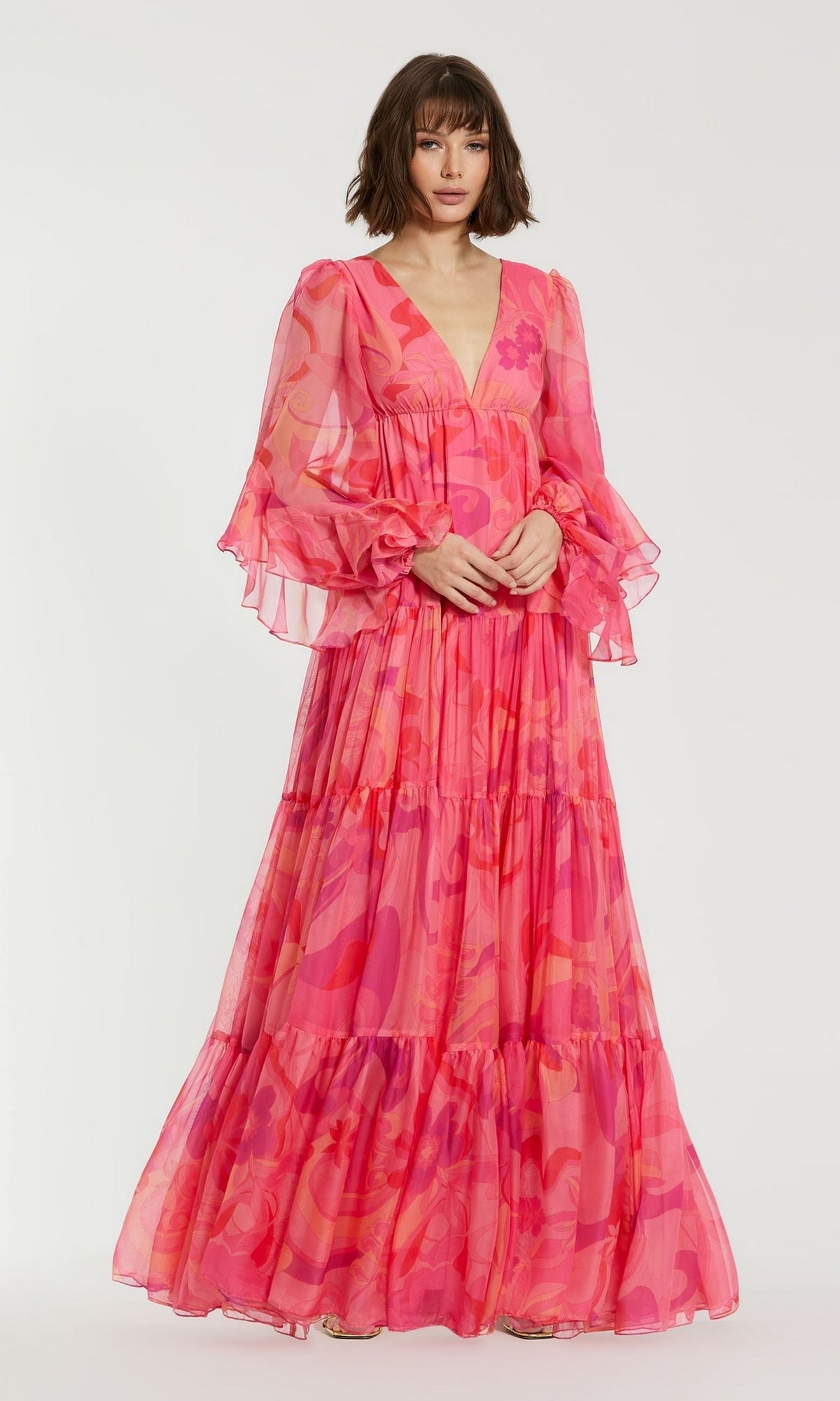 A woman wears the Long Formal Dress 68782 by Mac Duggal—a floor-length pink floral gown with sheer ruffled sleeves, deep V-neckline, and tiered A-line silhouette—posed against a plain white background.