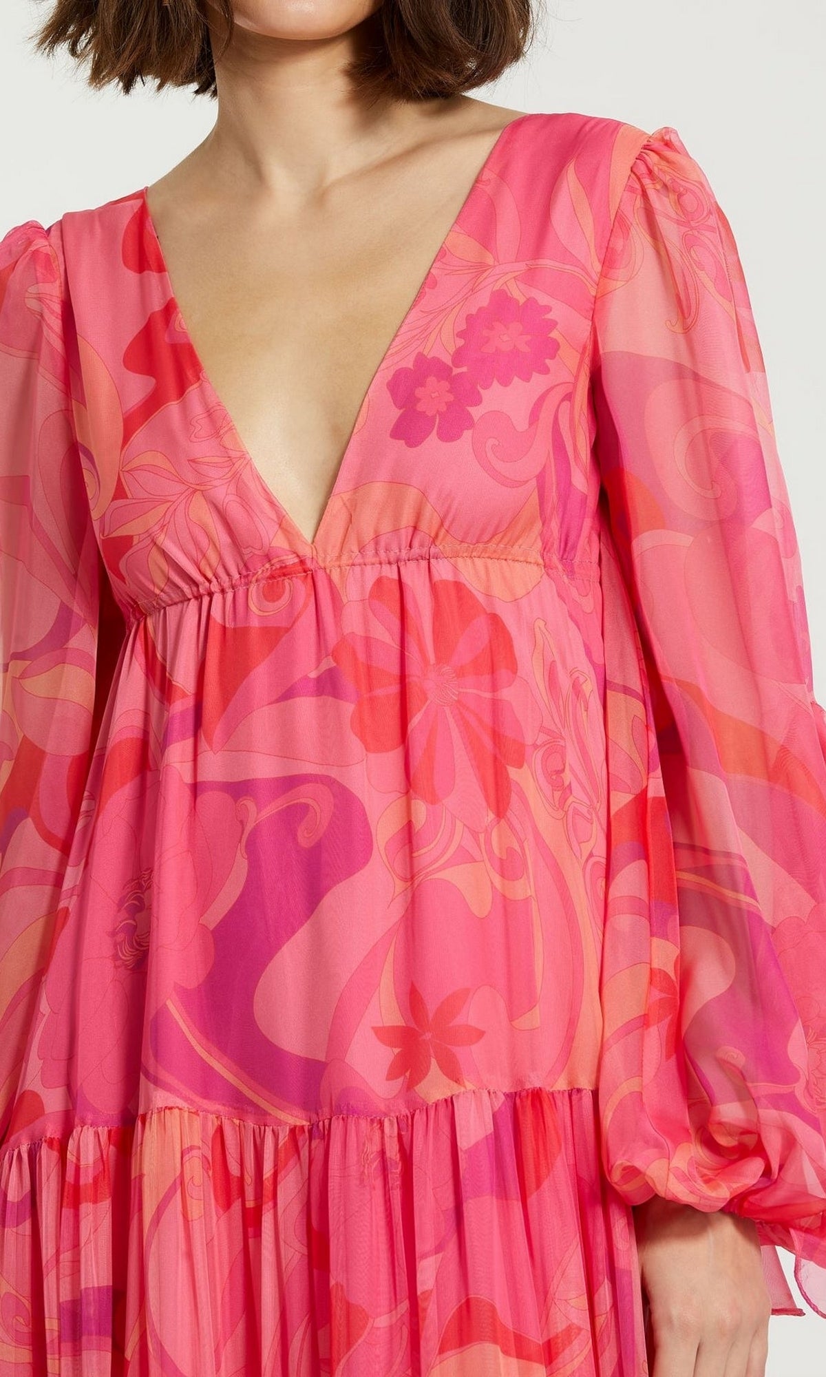 A woman wears the Long Formal Dress 68782 by Mac Duggal—a bright pink, long-sleeve, v-neck a-line tiered dress with a bold abstract floral pattern. Only the upper half of this vibrant style is visible.