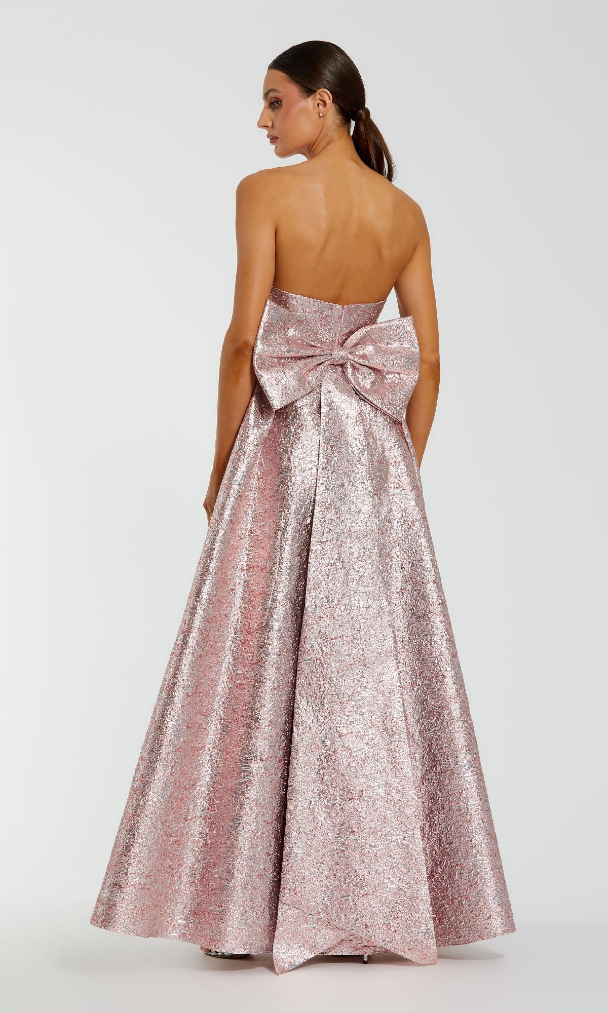 A woman models the Strapless Raspberry Formal Gown by Mac Duggal (68805), a long metallic pink dress with a back bow detail, standing against a plain background.