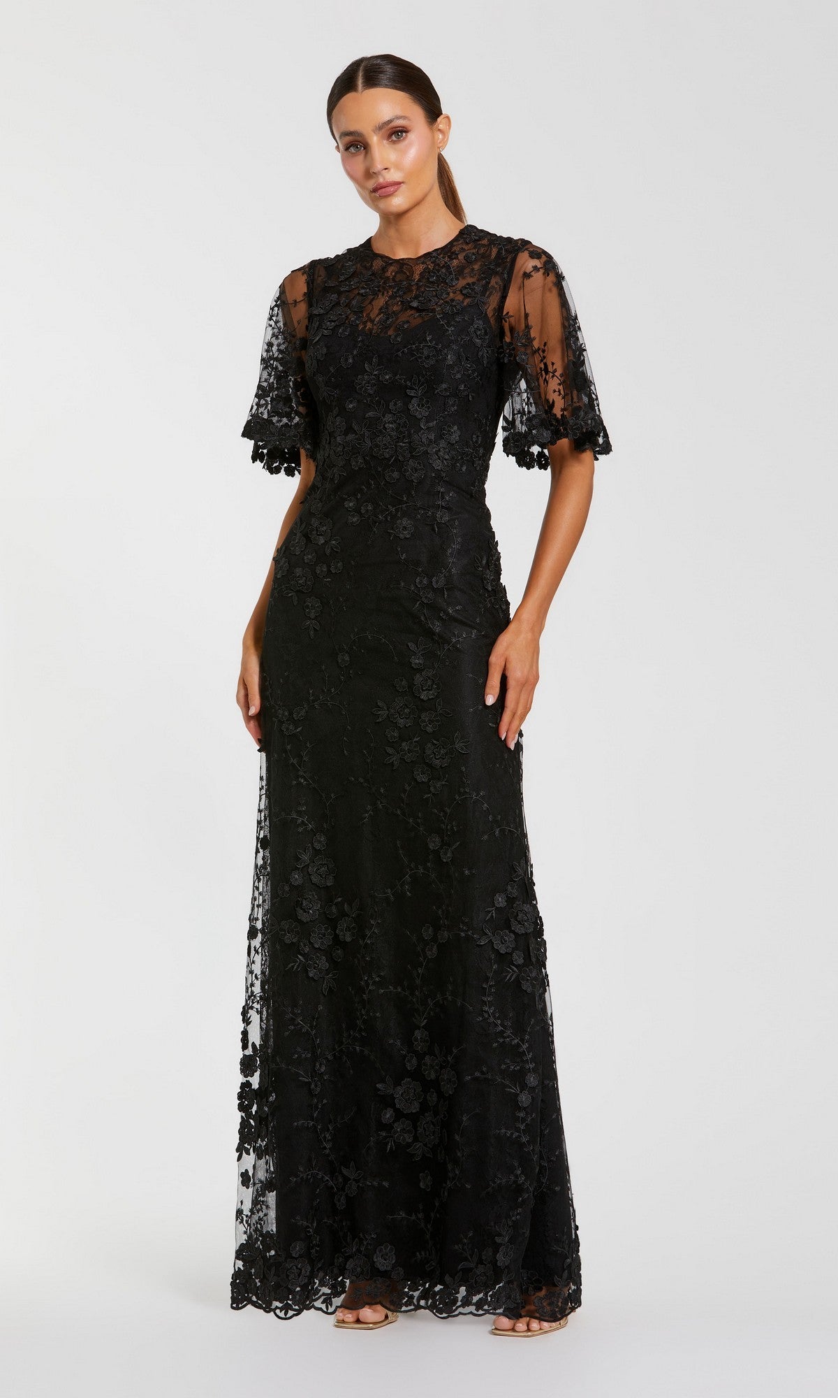 A woman wears the Flutter-Sleeve Long Formal Dress (Mac Duggal 68810), a floor-length black gown featuring short flutter sleeves, lace fabric, floral embroidery, and sheer details, set against a plain light background.