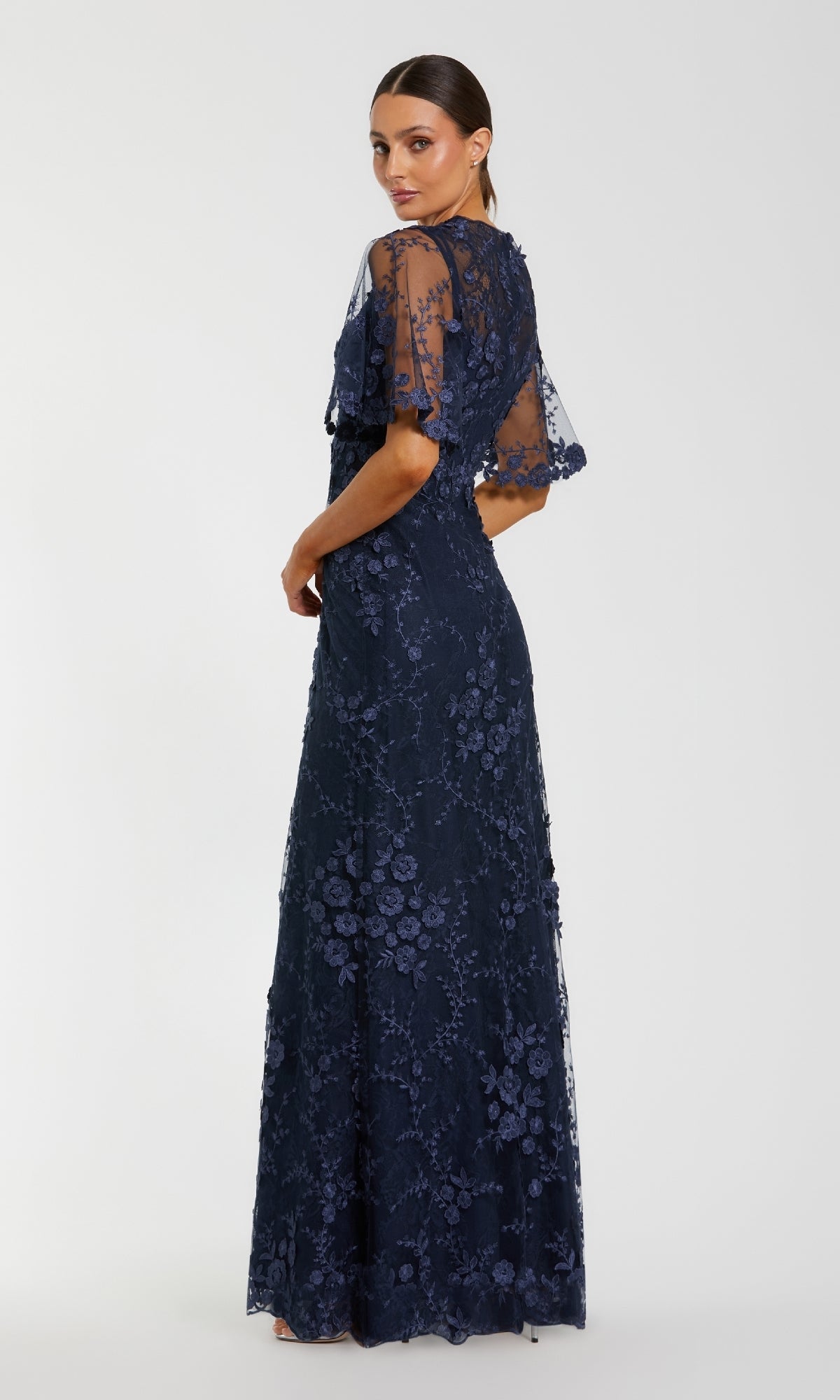 A woman in the Flutter-Sleeve Long Formal Dress by Mac Duggal (style 68810)—a navy blue, floor-length gown with floral lace and sheer short sleeves—stands turned slightly away from the camera against a plain light background.