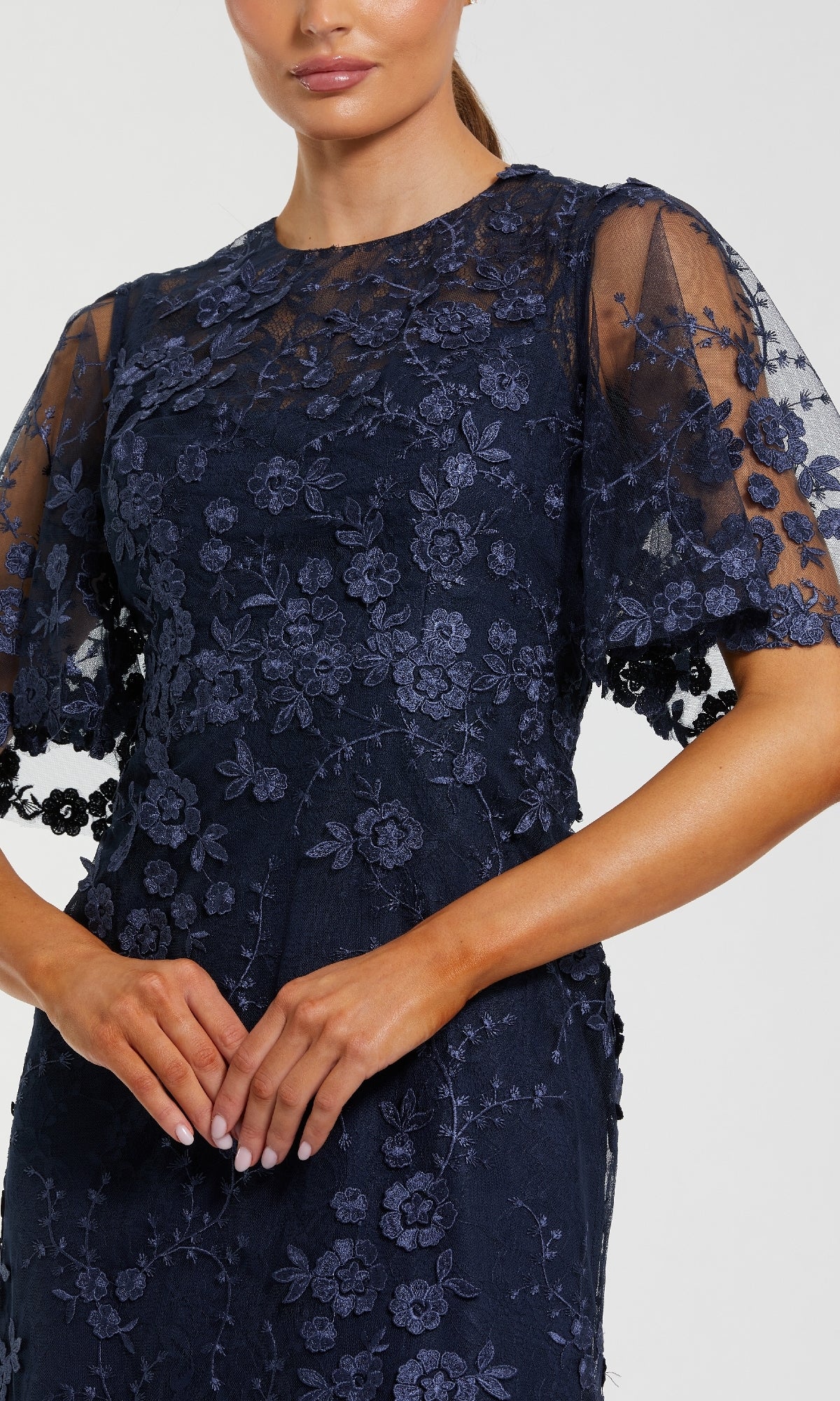 A woman stands with hands clasped, wearing the Mac Duggal 68810 Flutter-Sleeve Long Formal Dress in navy blue lace with floral embroidery and sheer short flutter sleeves.