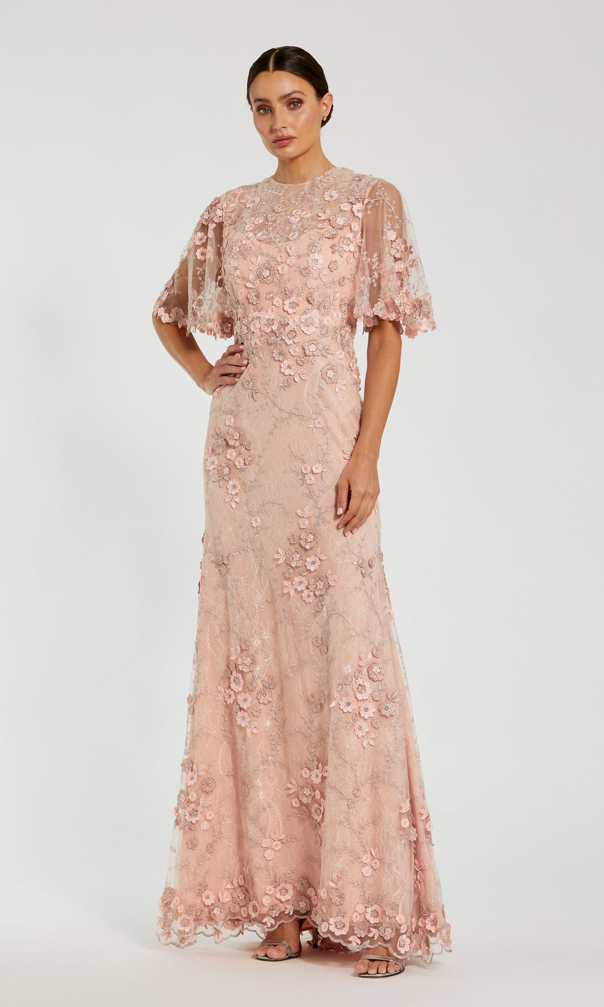 A woman stands against a white background wearing the Mac Duggal 68810 Flutter-Sleeve Long Formal Dress, a floor-length light pink lace gown with floral embroidery and short sheer flutter sleeves.