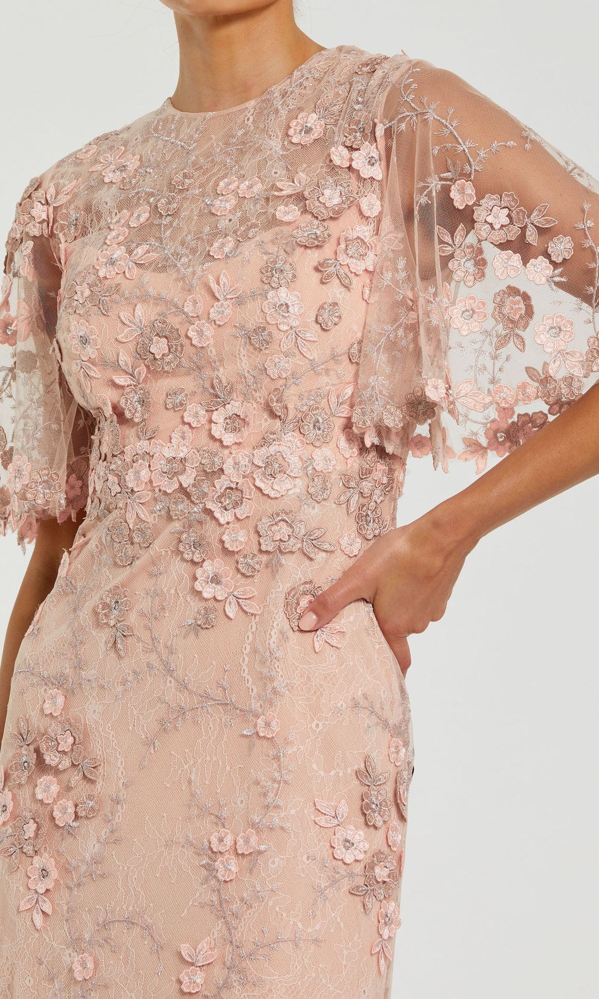 A model wears the Mac Duggal 68810 Flutter-Sleeve Long Formal Dress—a light pink gown with floral appliqué embroidery and sheer flutter sleeves, shown from neck to mid-thigh.