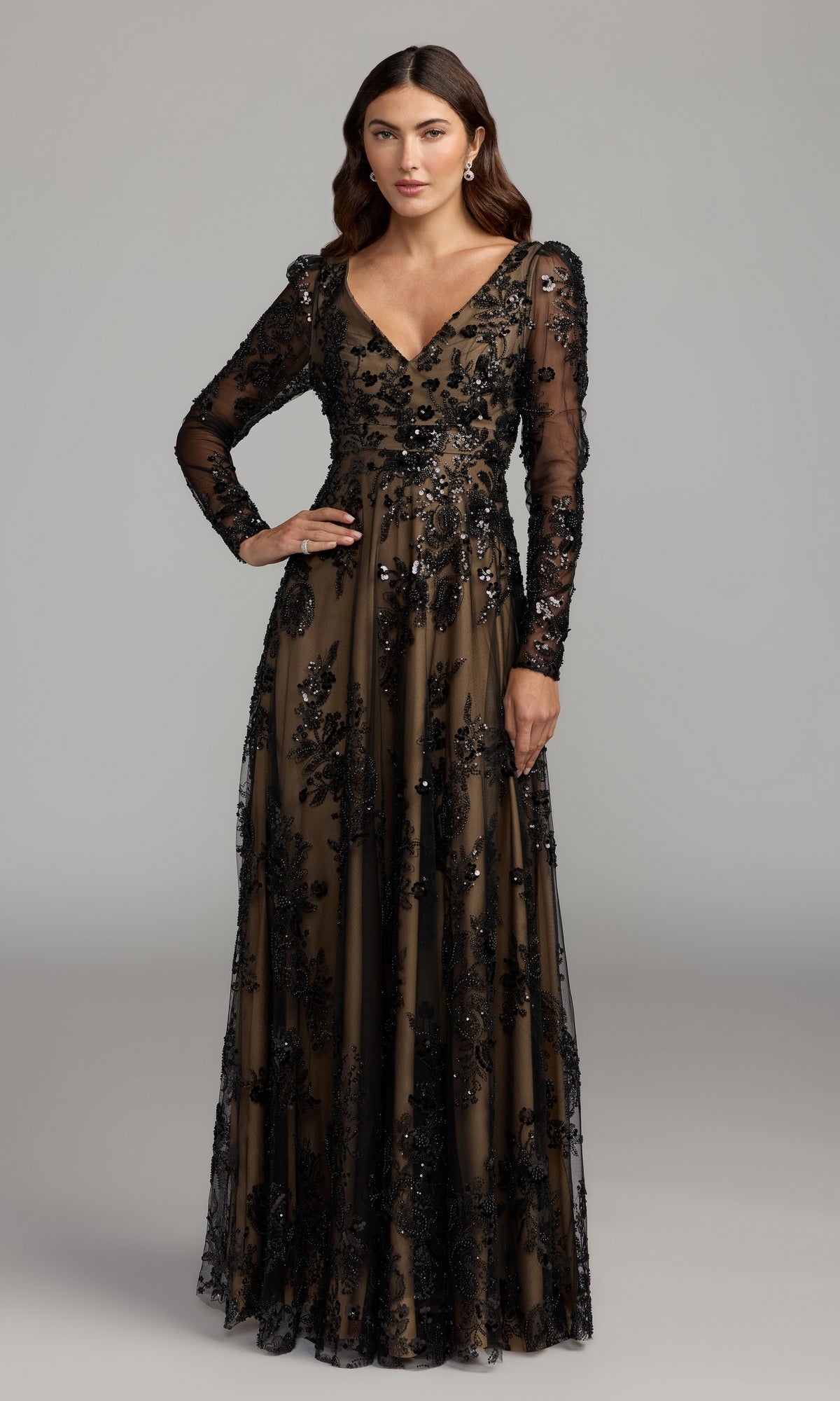 A woman stands against a plain gray background wearing the Mac Duggal 68908, a sophisticated long sleeve formal dress in black nude, featuring sheer fabric and sequin floral embellishments—ideal as an embroidered gown for the mother of the bride or any elegant occasion.