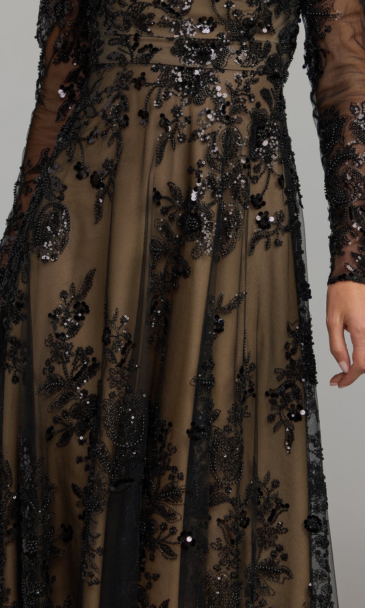 A close-up of a person wearing the Mac Duggal 68908 Long Sleeve Long Formal Dress, featuring a sheer black fabric adorned with intricate bead and sequin floral embroidery, highlighting part of the arm and skirt.