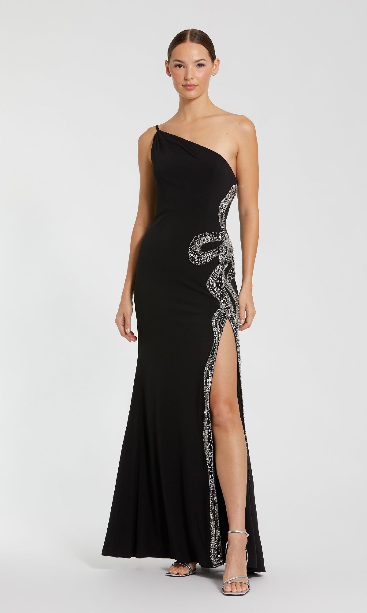 A woman in the Mac Duggal 68932 long formal dress—black, one-shoulder, with silver embellishments and a thigh-high slit—stands against a plain background, embodying timeless elegance.