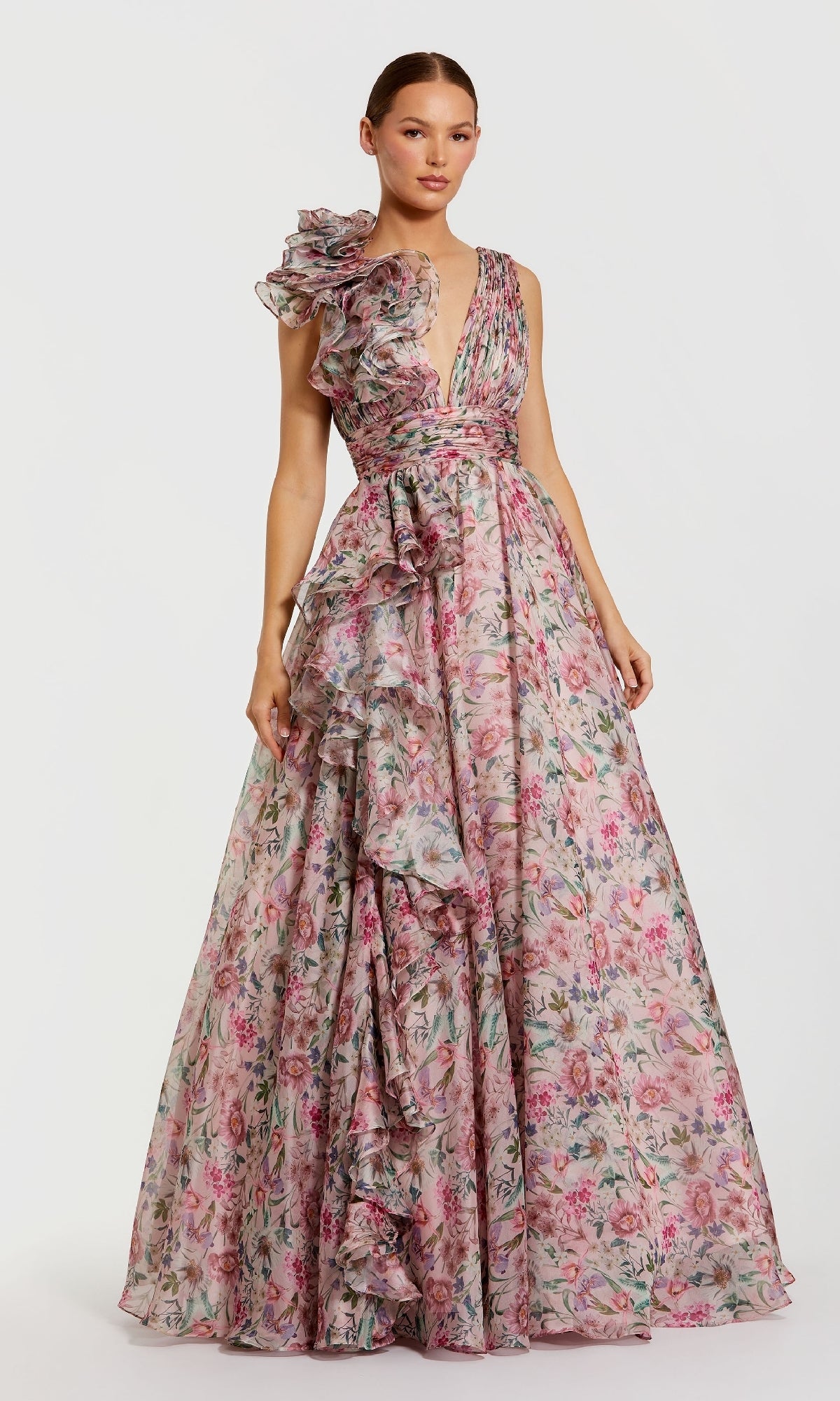 A woman wears the Mac Duggal 68947: a floor-length, sleeveless formal dress with a pink floral print, deep V-neckline, and asymmetric ruffled details on the bodice and skirt.