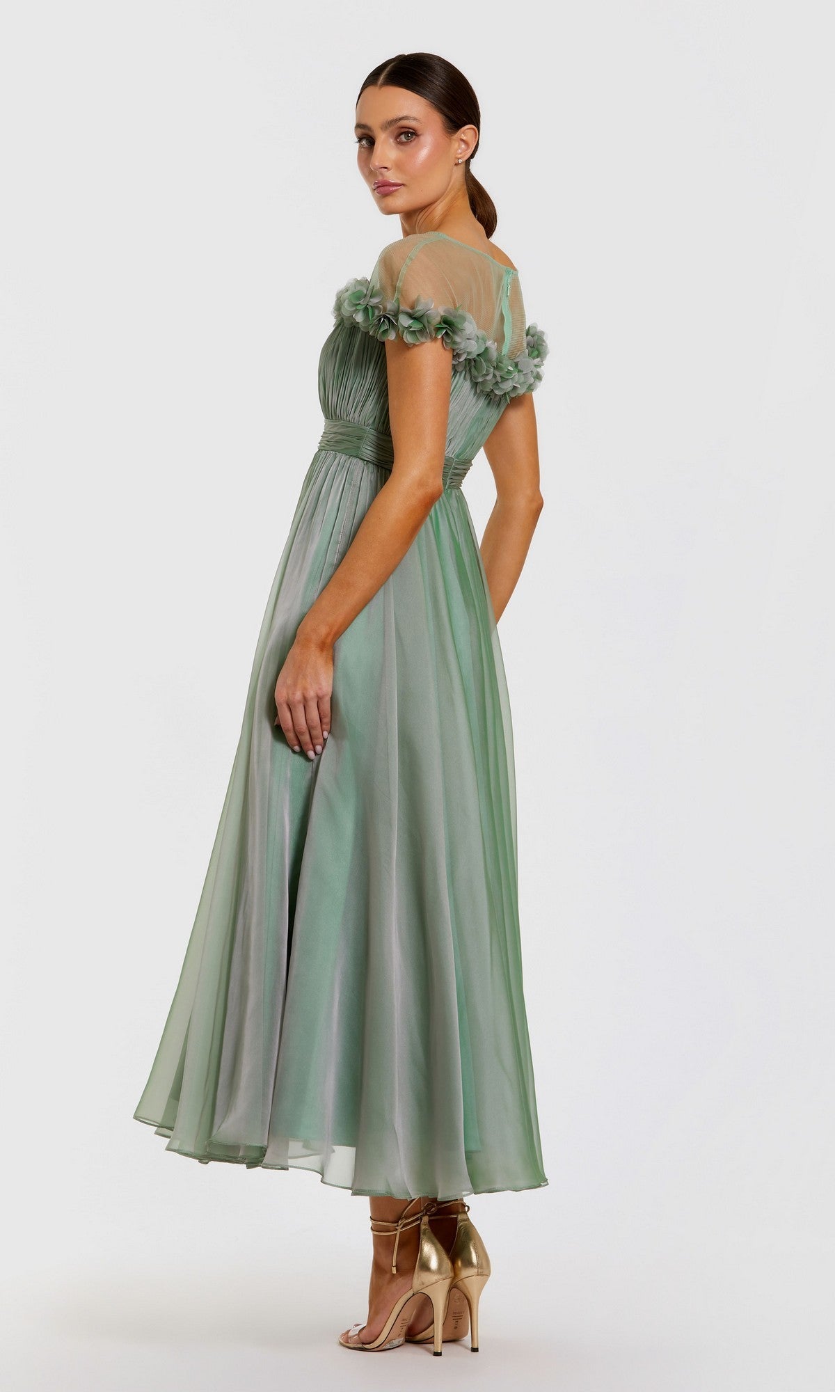 A woman models the Mac Duggal 68966 Tea-Length Party Dress—a light green, off-the-shoulder fit-and-flare design featuring tulle details and floral accents—paired with gold high-heeled sandals against a plain light background.