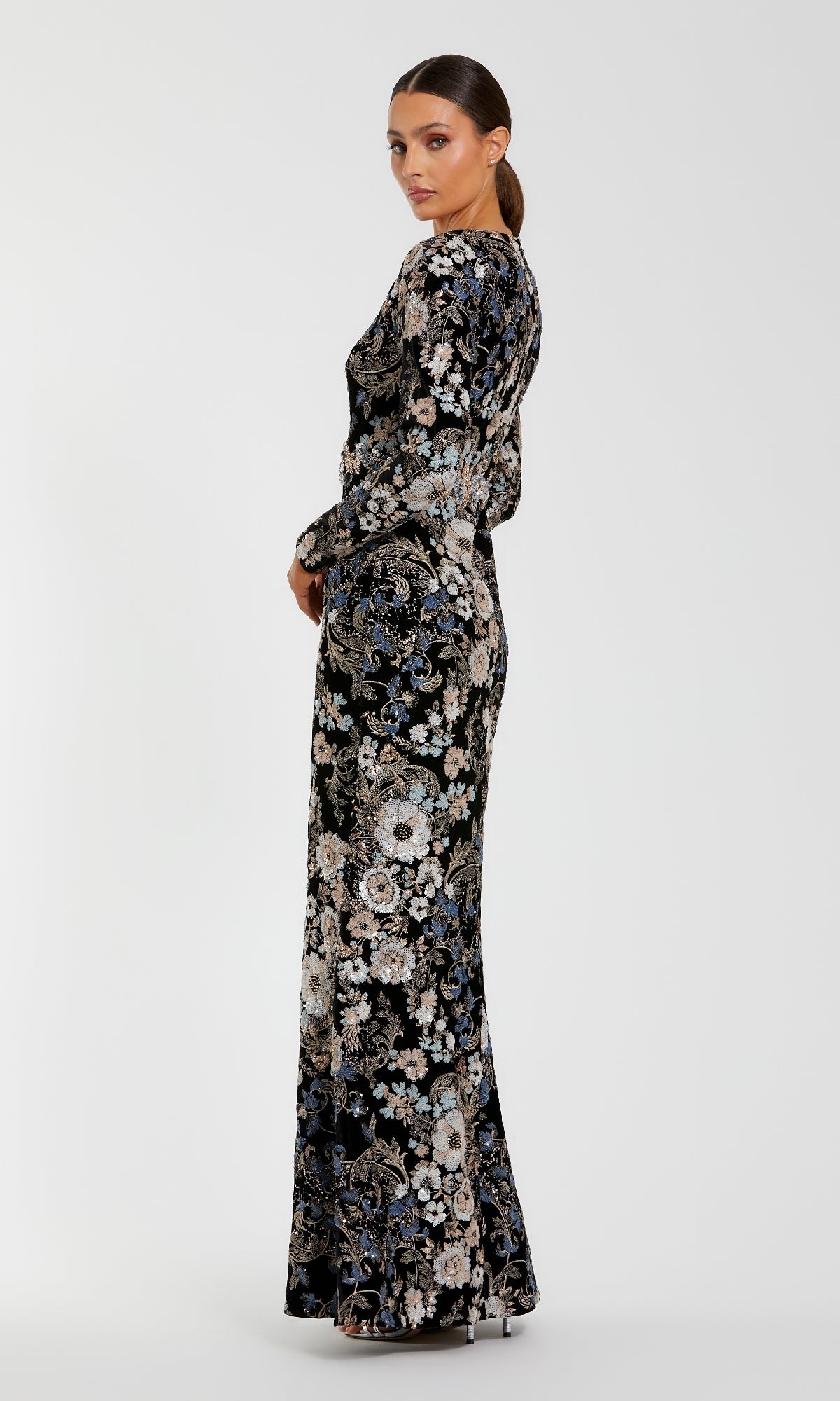 Woman wearing the Mac Duggal 68978 Black Velvet Print Formal Gown with long sleeves, floor length, embroidered sequin floral print, and side slit, standing sideways against a plain white background.