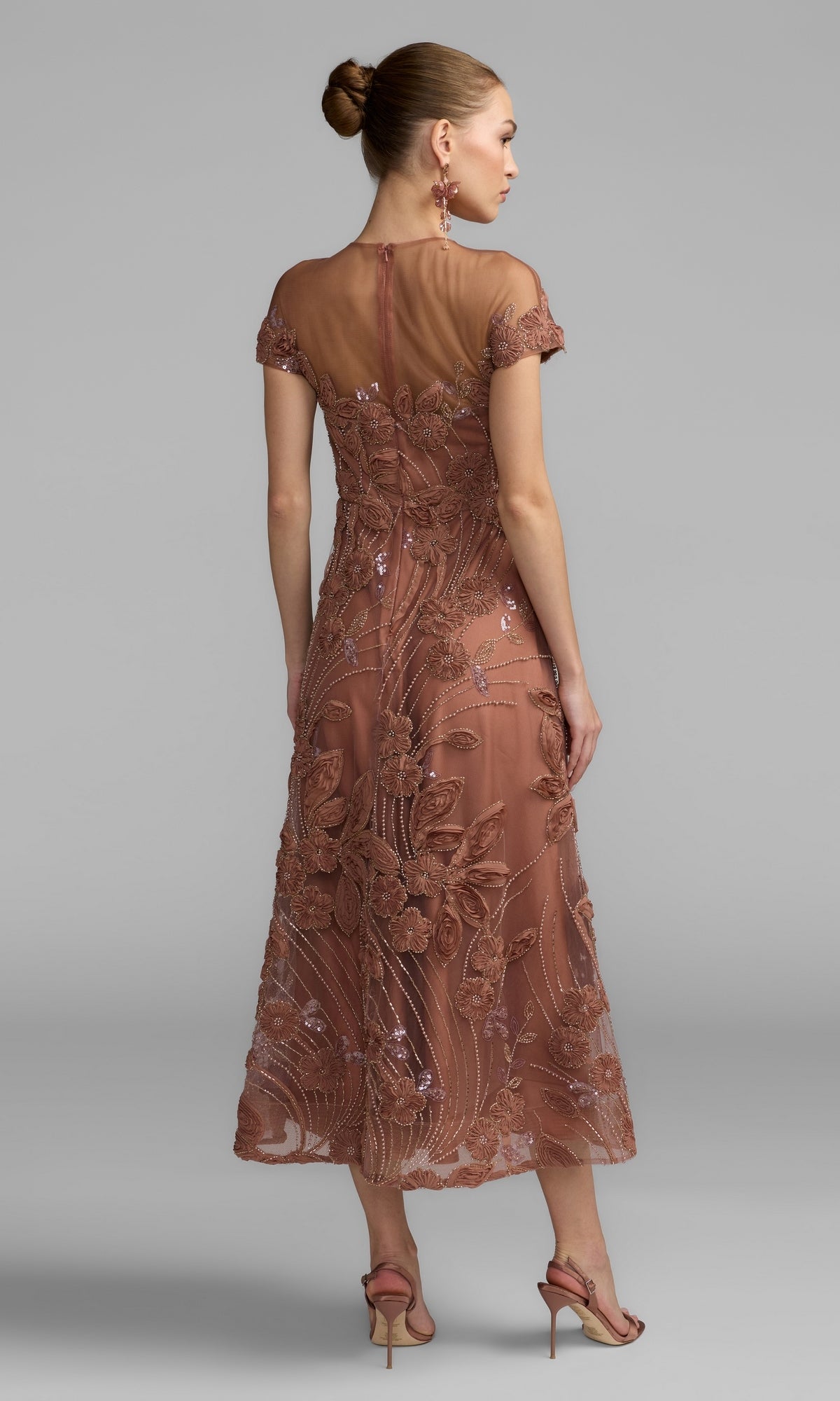 A woman stands against a plain gray background, wearing a cap-sleeve black formal dress—specifically the Mac Duggal 69003—with elegant high-heeled sandals. The dress features an embroidered design, sheer back, and floral patterns that exude timeless romantic elegance.