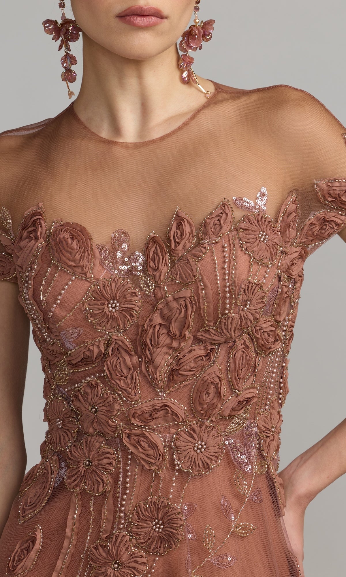 A woman is shown from shoulders to waist wearing a sheer, rose-colored dress adorned with intricate floral embroidery and matching floral earrings—a striking contrast to the classic Mac Duggal 69003 Cap-Sleeve Black Formal Dress featuring elegant beaded lace.