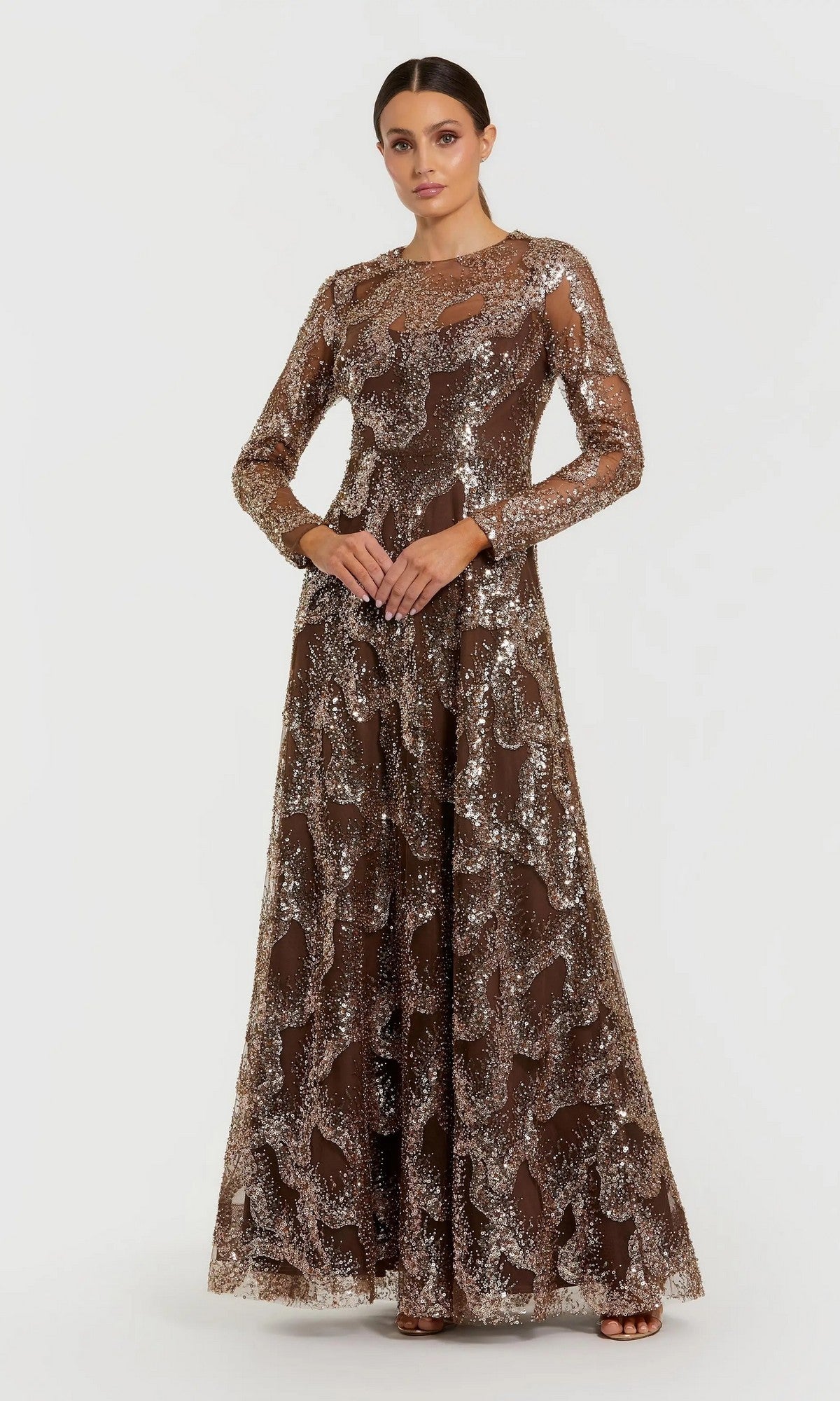 A woman stands against a plain white background, hands clasped in front and wearing a neutral expression. She is dressed in the Mac Duggal 69005 long formal dress—an elegant, full-length gown with modest long sleeves and shimmering silver sequin patterns accenting the rich brown fabric.