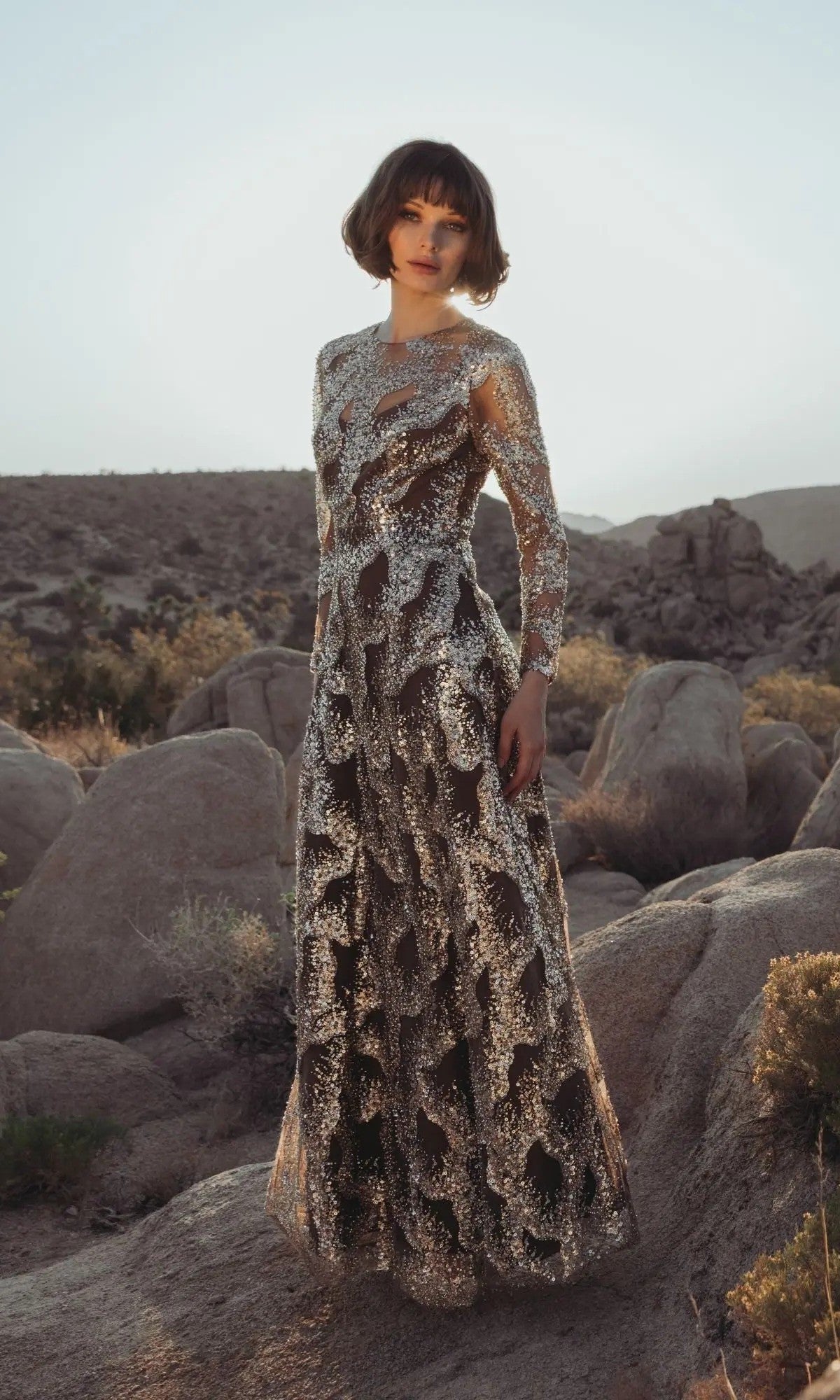 A person wearing the bronze Mac Duggal 69005 long formal dress stands on rocky terrain outdoors, with short brown hair and a neutral expression, surrounded by desert plants and boulders under a clear sky.