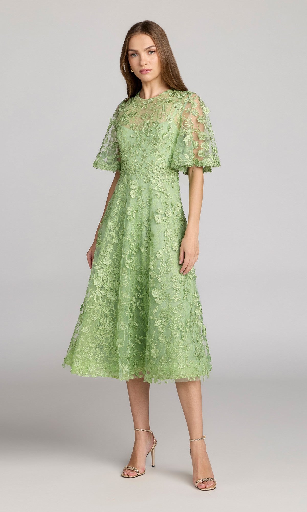 A woman wears the Mac Duggal 69036 Lace Midi Wedding Guest Dress, featuring light green lace with floral details and short sleeves, paired with open-toe heels against a plain gray background—perfect for wedding guests.