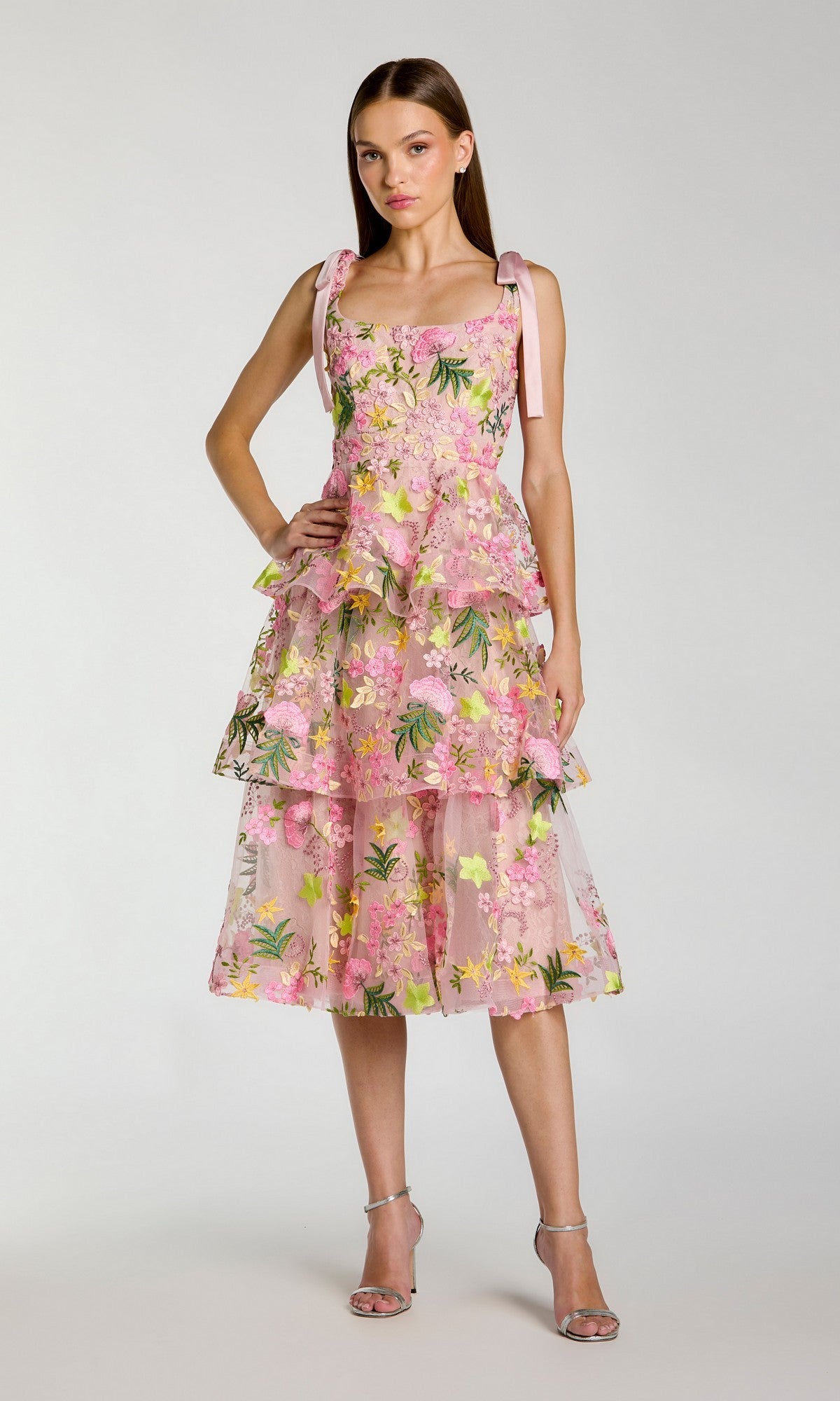 A woman in the Mac Duggal 69043 Midi Party Dress in Blush Multi—featuring a sleeveless, layered floral design with a tiered A-line skirt and delicate shoulder ties—paired with silver high-heeled sandals, stands against a plain light background.