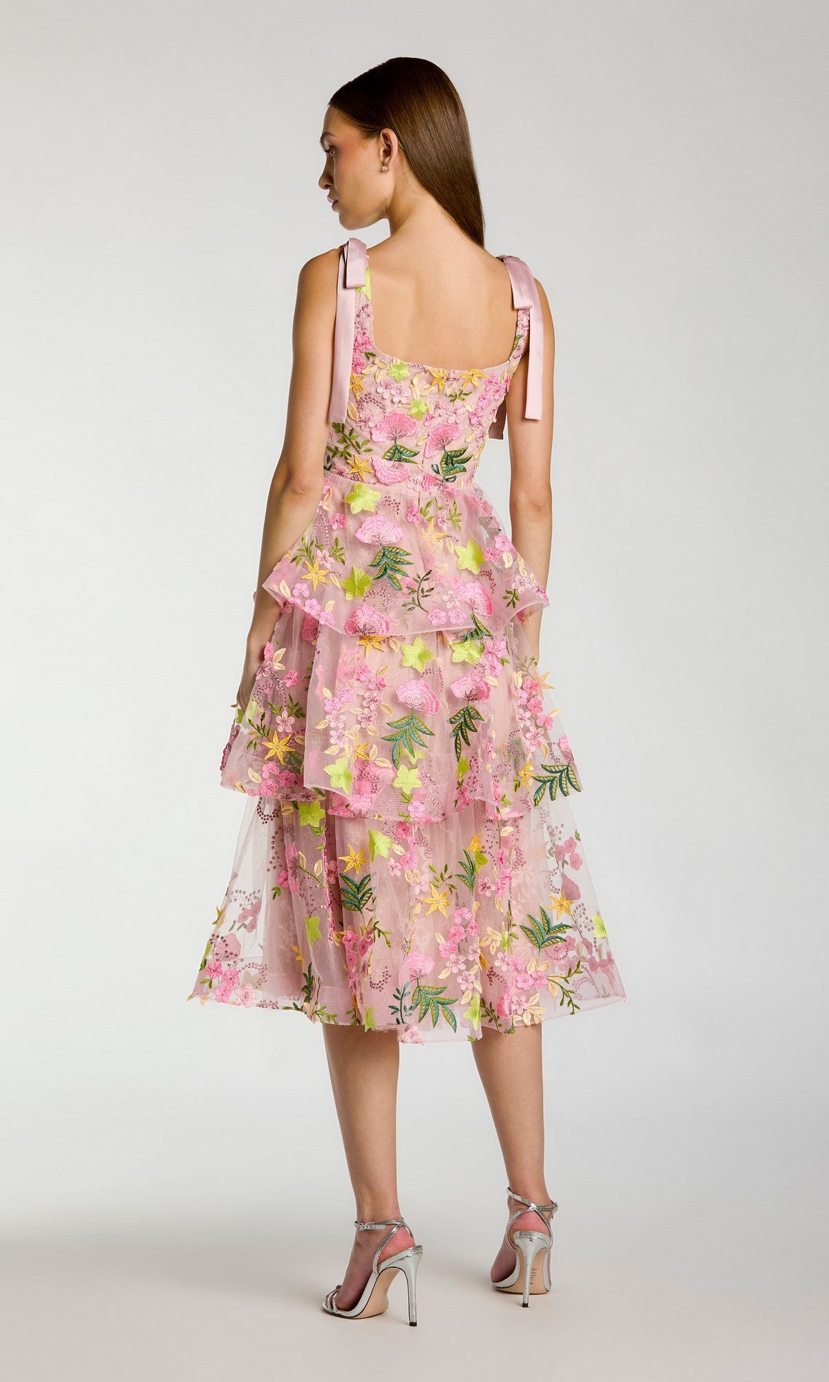 A woman stands against a plain light background, facing away, wearing the Mac Duggal 69043 Midi Party Dress—a pink floral wedding guest dress featuring ribbon straps and a tiered A-line skirt—paired with silver high-heeled sandals.