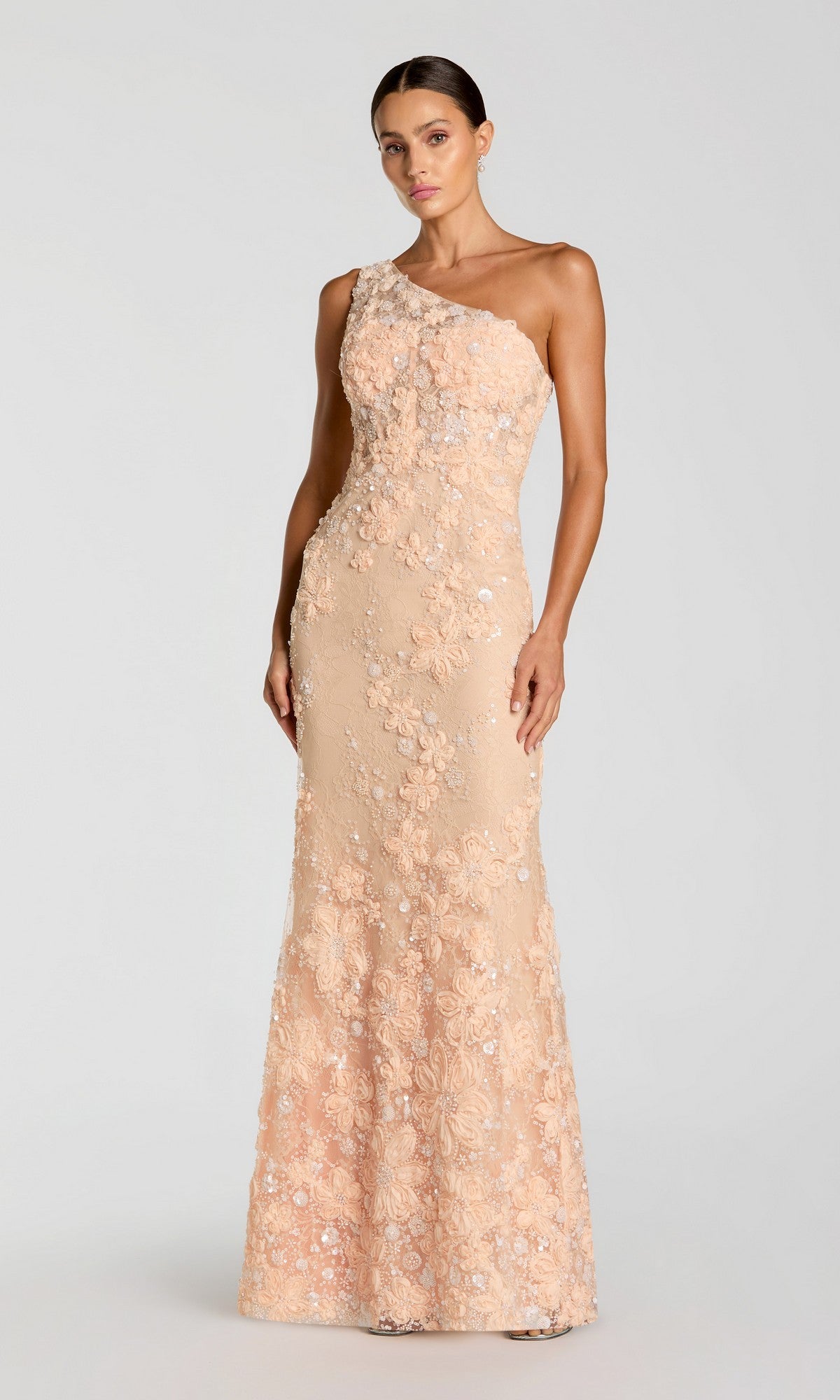 A woman wearing the Mac Duggal 69052 long formal dress in peach—an elegant one-shoulder gown adorned with intricate sequin floral appliqué—stands against a plain light background.