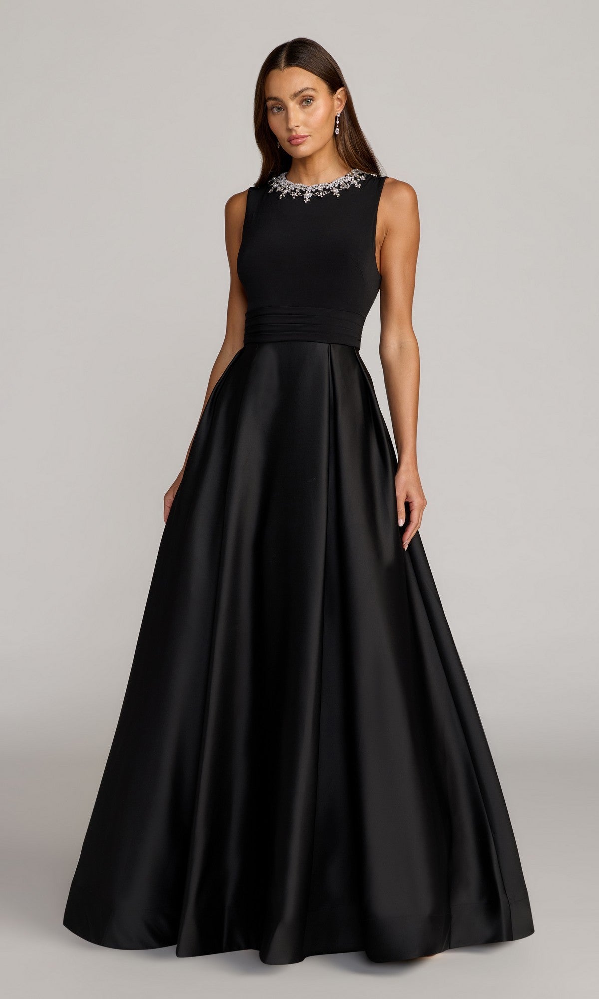 A woman wearing the Mac Duggal 69084 long formal dress in black, featuring a sleeveless design, full skirt, and jeweled neckline, stands against a plain light background.