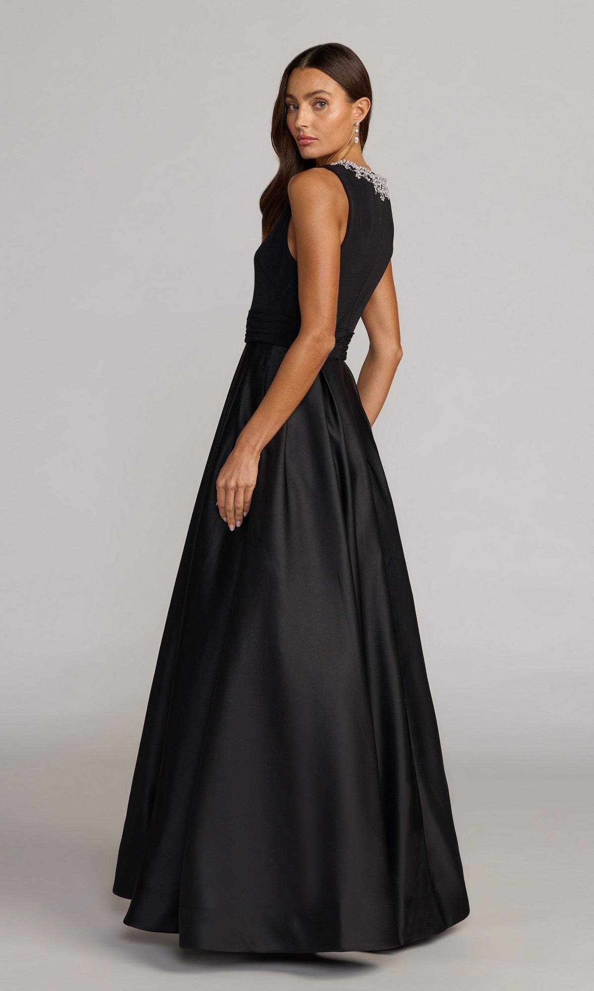 A woman stands against a plain background, looking over her shoulder in the Long Formal Dress: Mac Duggal 69084—a sleeveless black gown with a jeweled neckline and a classic elegant silhouette.