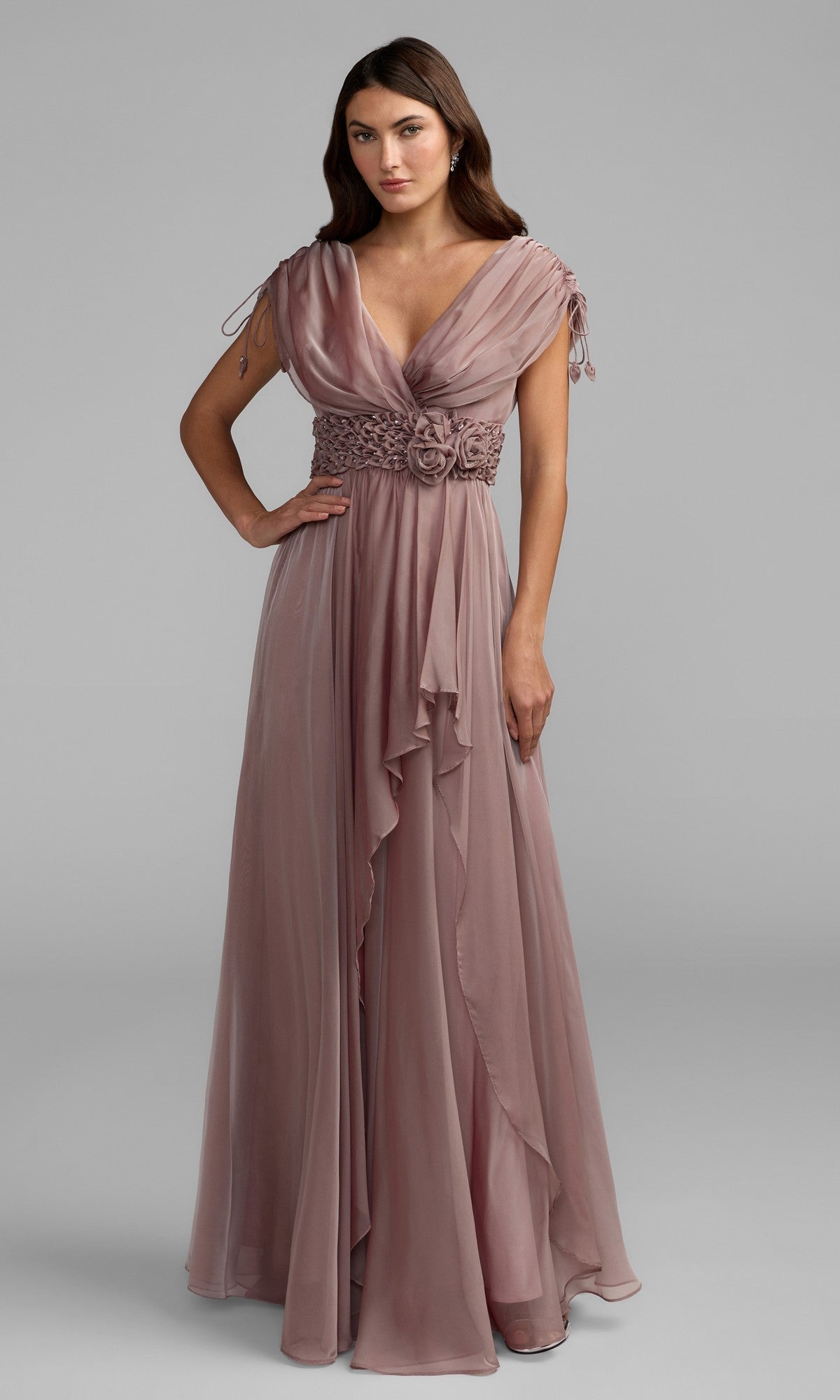 A woman wearing the Long Formal Dress: Mac Duggal 69089 in mocha—a floor-length chiffon formal gown featuring ruffled accents, a V-neckline, cap sleeves with ties, and a crystal-embellished waistband—stands against a neutral gray background.