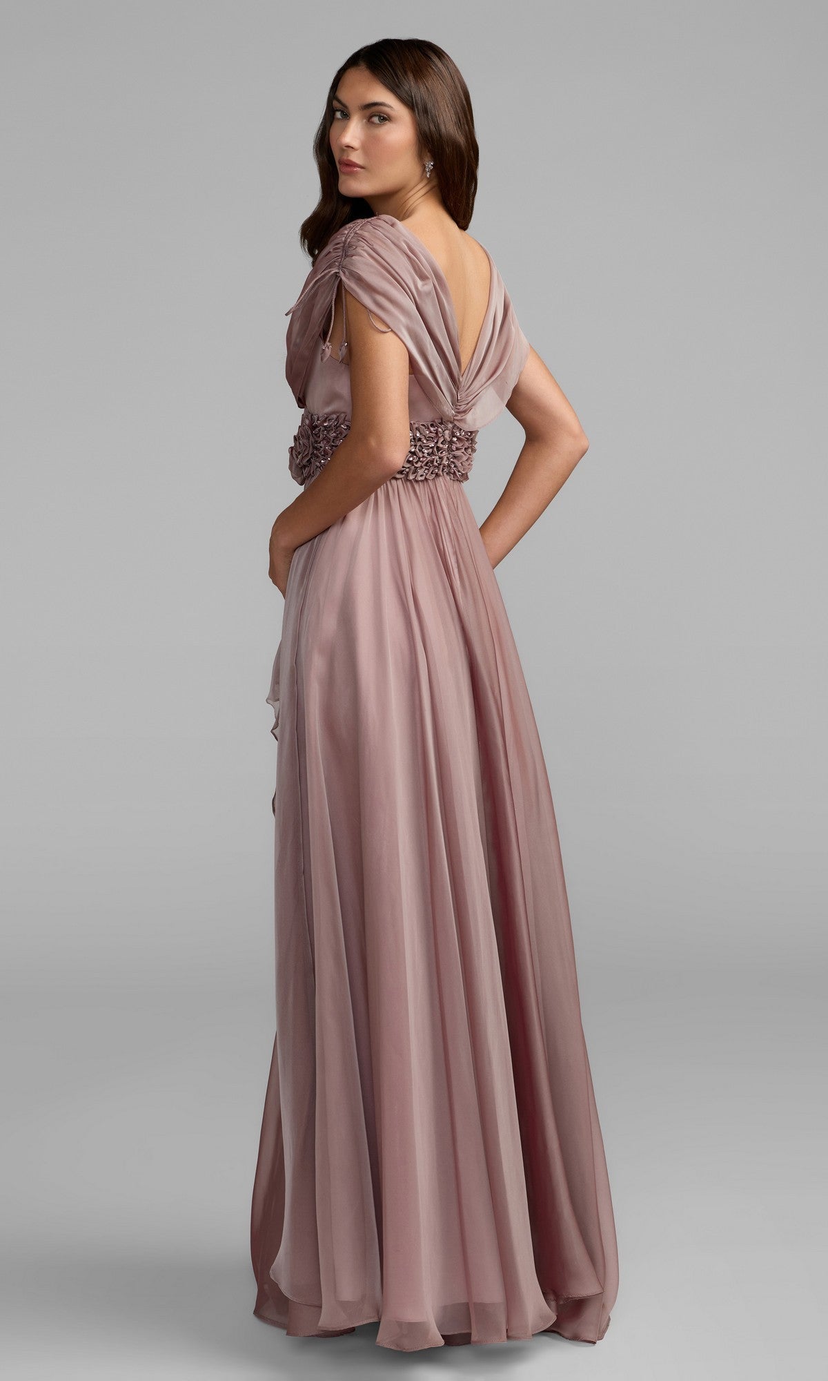 A woman models the Mac Duggal 69089 long formal dress in mauve chiffon, featuring draped sleeves and a crystal-embellished waistband, as she stands against a plain gray background and faces slightly away from the camera.