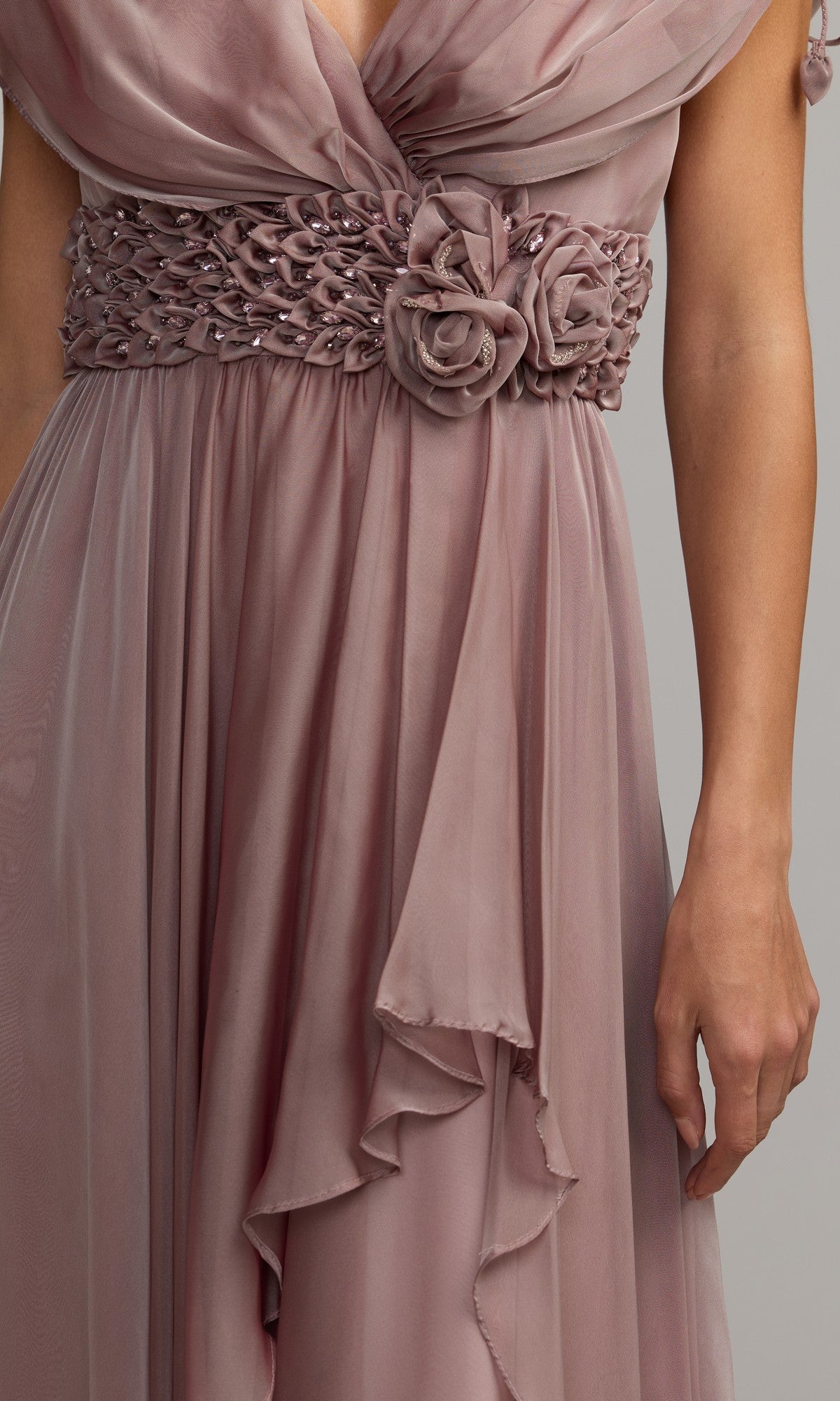 A close-up of the Long Formal Dress: Mac Duggal 69089 in mauve chiffon, showcasing its ruched bodice, crystal-embellished waistband with delicate floral detailing, and sheer flowing fabric, worn by a person with one arm visible.