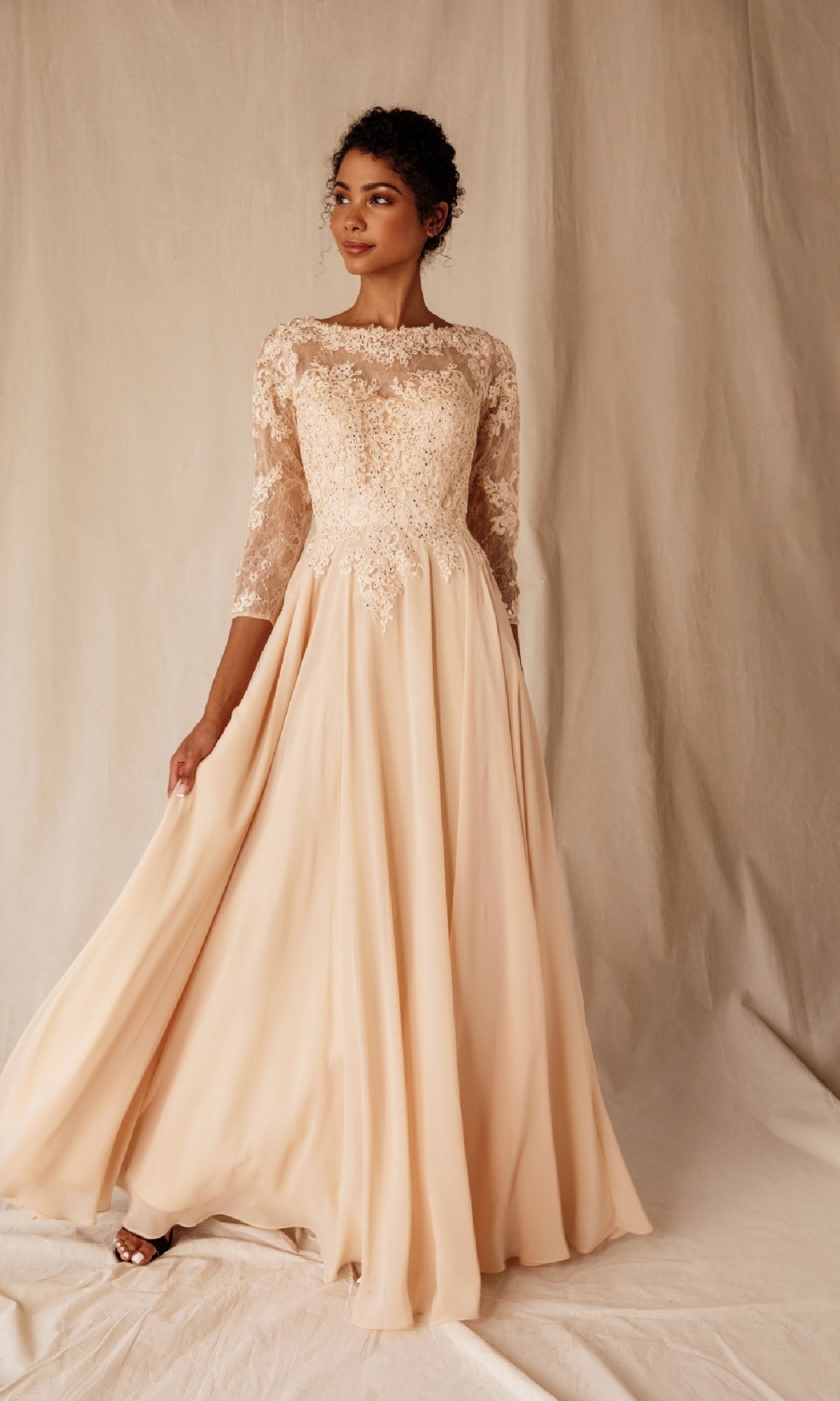A woman poses before a neutral backdrop in the Eureka 6909 long-sleeve formal dress, showcasing its modest A-line silhouette with an ivory lace bodice and flowing chiffon skirt, holding one side of the gown.