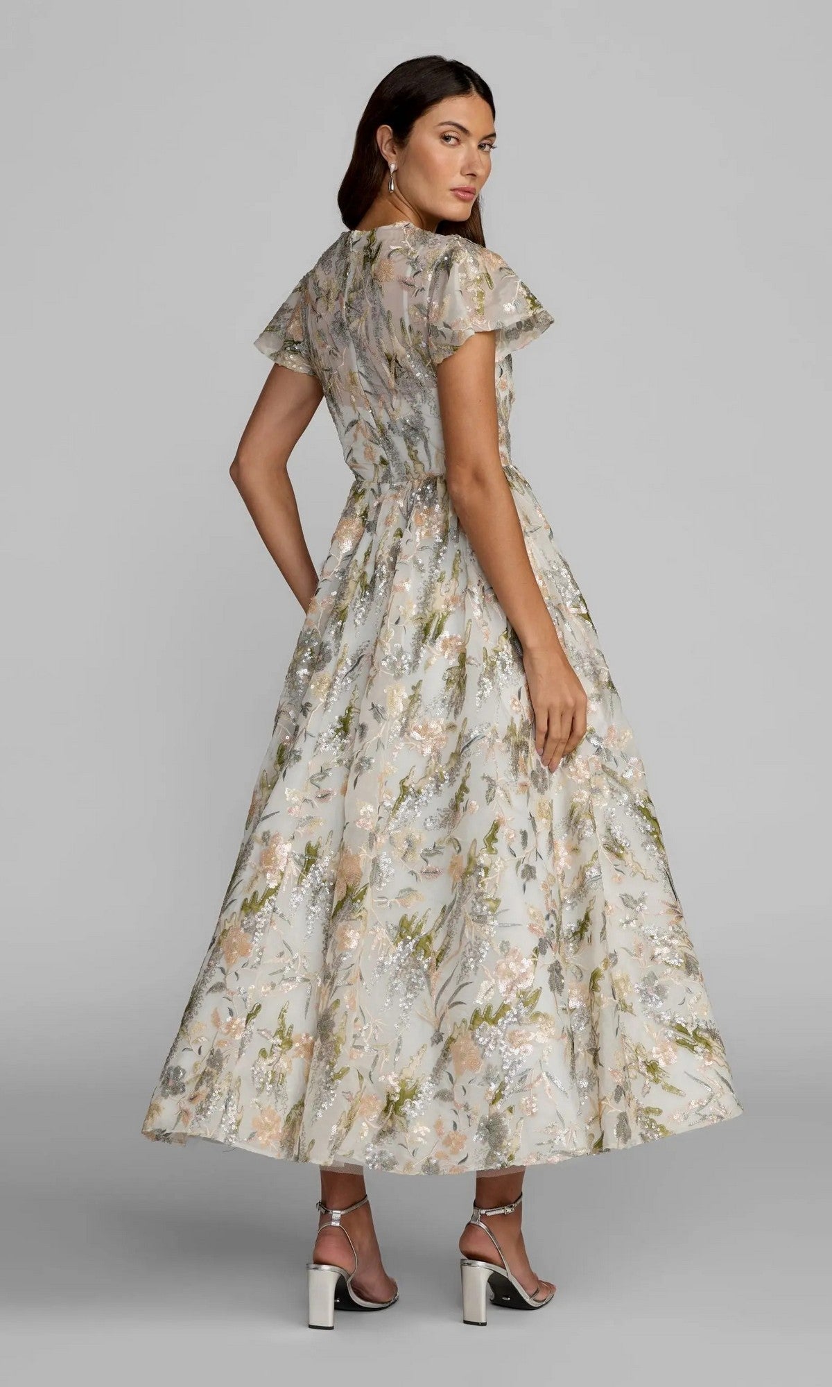 A woman stands sideways in the Mac Duggal 69107 Tea-Length Party Dress—a modest, floral design with short sleeves and an ankle-length hem—paired with silver high heels, set against a plain background.