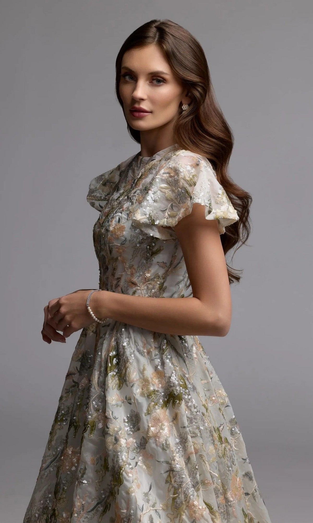A woman stands with hands clasped, gazing at the camera while wearing the Mac Duggal 69107 Tea-Length Party Dress—featuring a sequin floral pattern and flutter sleeves—set against a plain gray background.