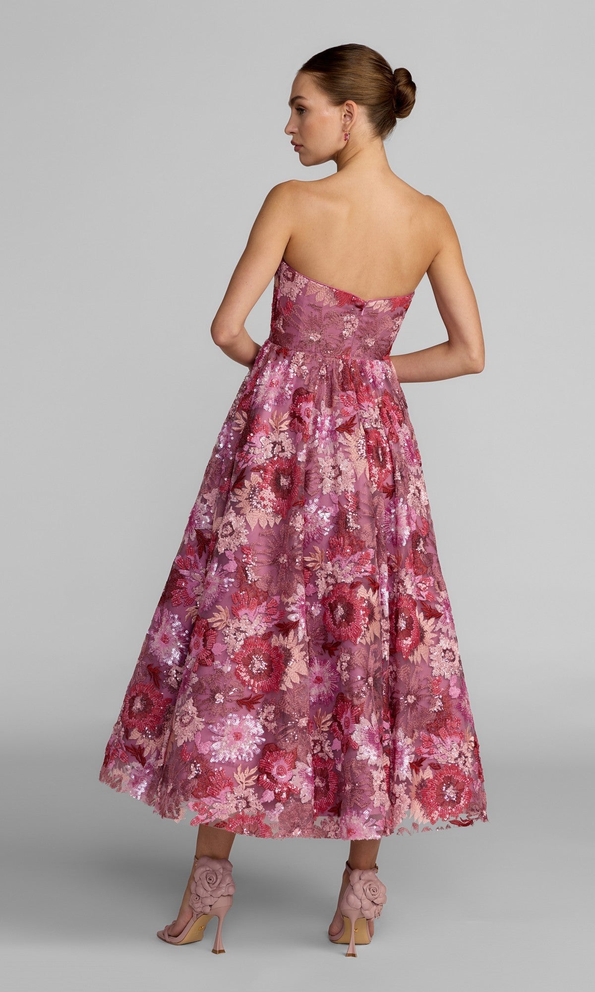 A woman in the Mac Duggal 69109 Tea-Length Party Dress, a pink strapless midi cocktail dress adorned with delicate floral sequin embroidery, stands facing away with her hair in a bun and matching high-heeled shoes.