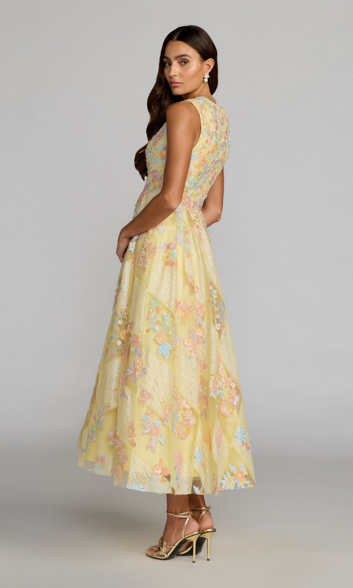 A woman stands in profile against a plain background, wearing the Tea-Length Party Dress: Mac Duggal 69111—an ankle-length, sleeveless yellow gown adorned with pastel floral embroidery—paired with gold heels and drop earrings.