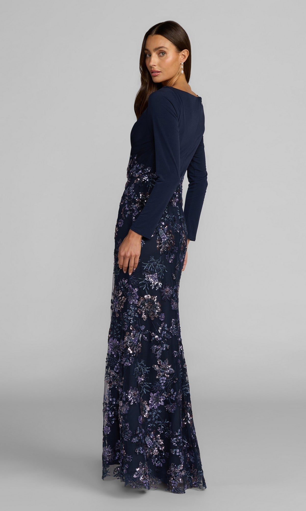 A woman faces away, modeling the Mac Duggal 69122 long formal dress, which features a long-sleeve navy top and a floor-length navy floral skirt, as she stands against a plain gray background.