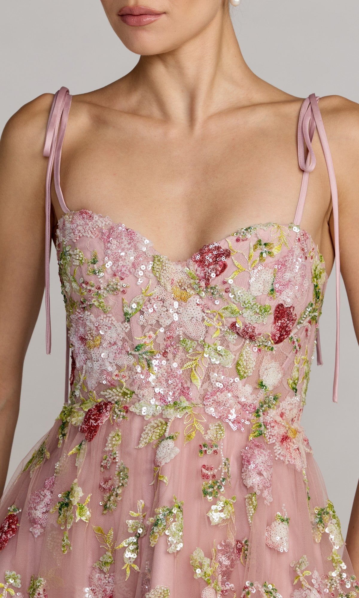A woman wearing the Long Formal Dress: Mac Duggal 69143, a pink floral gown adorned with delicate embroidery, sequin details, and thin shoulder straps tied in bows.