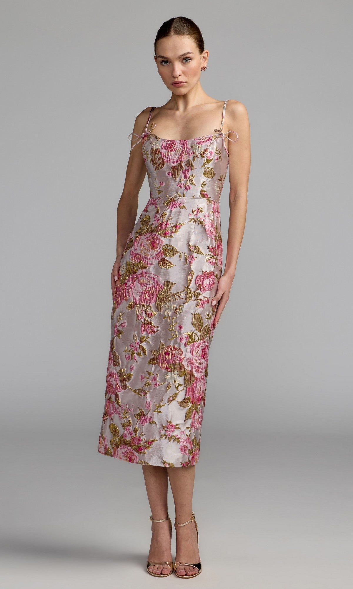 A woman wearing the Mac Duggal 69153 Midi Party Dress in Pink Multi, featuring spaghetti straps and a sleeveless design with pink and gold floral patterns, stands against a plain gray backdrop paired with high heels.