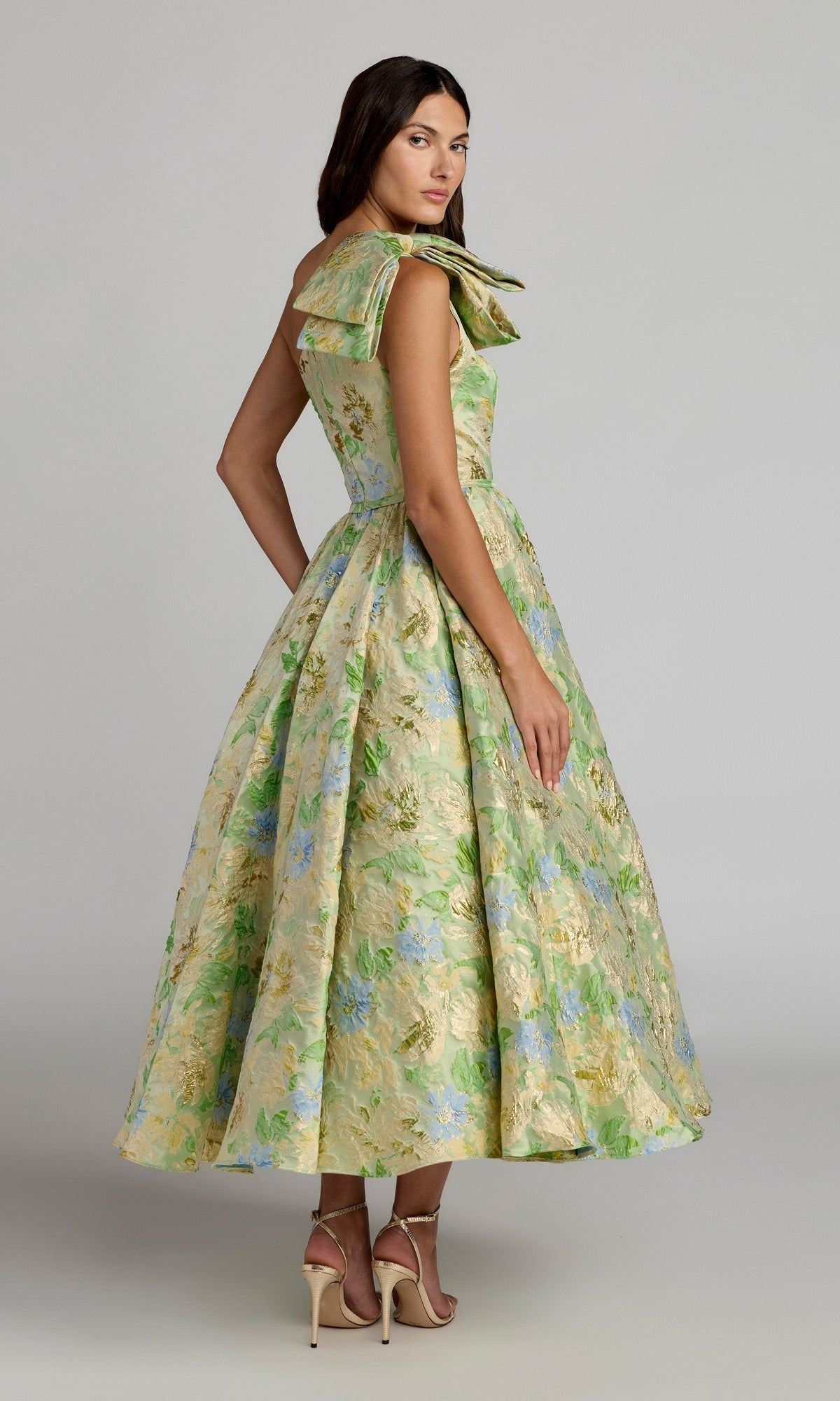A woman wears the Mac Duggal 69160 Tea-Length Party Dress, featuring a floral pattern, full skirt, and bow detail at the shoulder, styled with neutral heels against a plain background.
