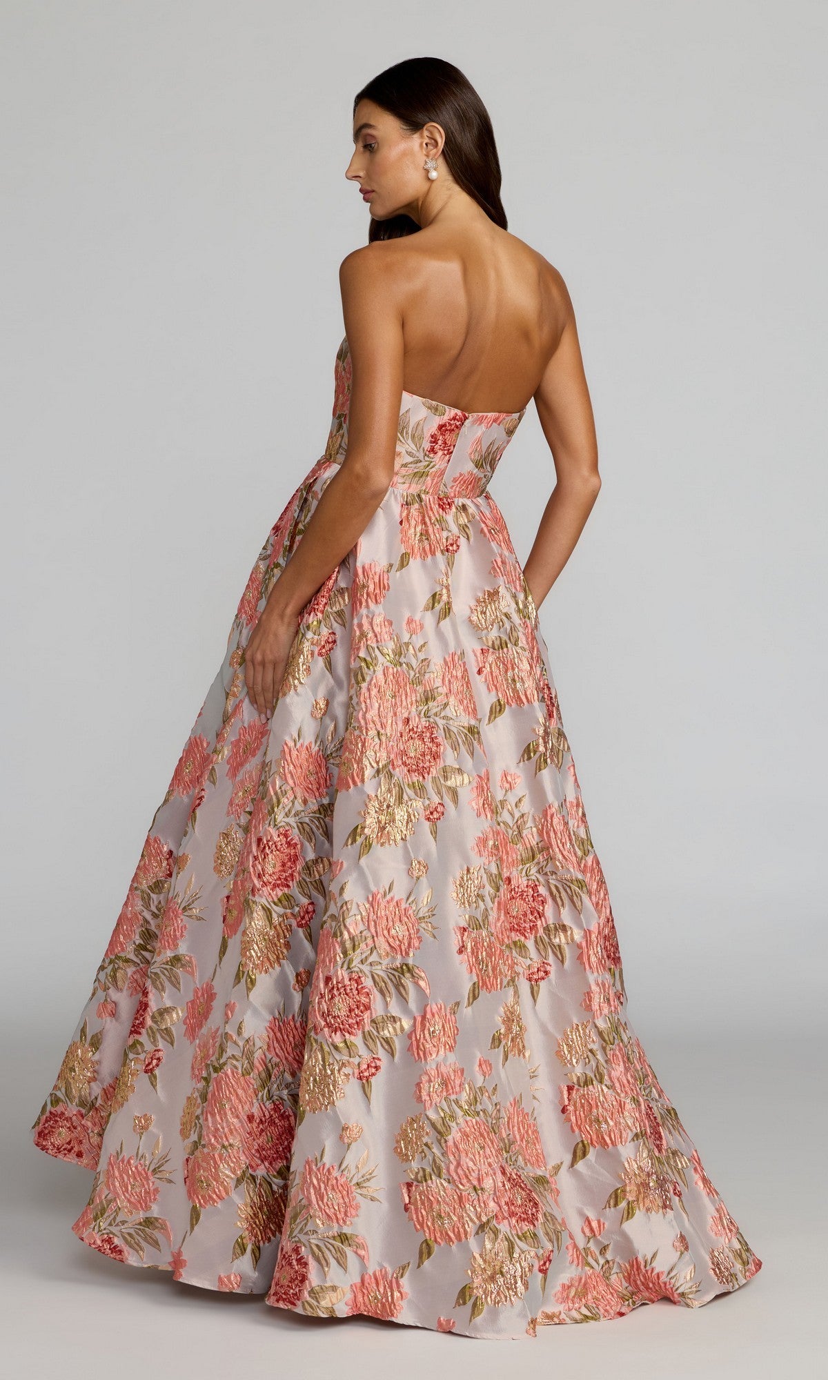 A woman wearing the Long Formal Dress: Mac Duggal 69169, a strapless A-line floor-length gown in pink and gold floral brocade, stands and looks over her shoulder.