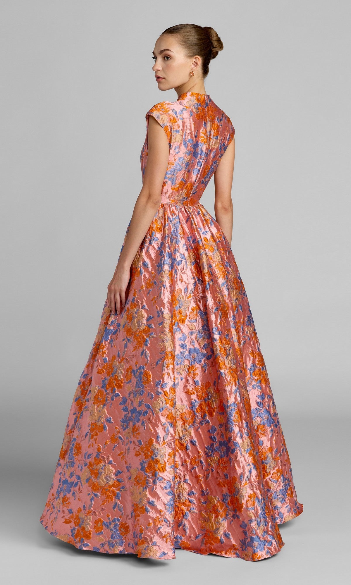 A woman stands against a plain gray background, wearing the Mac Duggal 69171 long formal dress—a floor-length, sleeveless gown adorned with intricate floral brocade in vibrant pink, orange, blue, and gold hues.