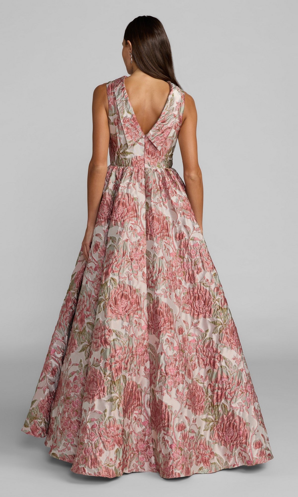 A woman is seen from behind wearing the Mac Duggal 69177 long formal dress, a sleeveless, floor-length ballgown crafted from pink floral brocade with a deep V-back. The gown features a stunning blend of pink, green, and beige patterned fabric.