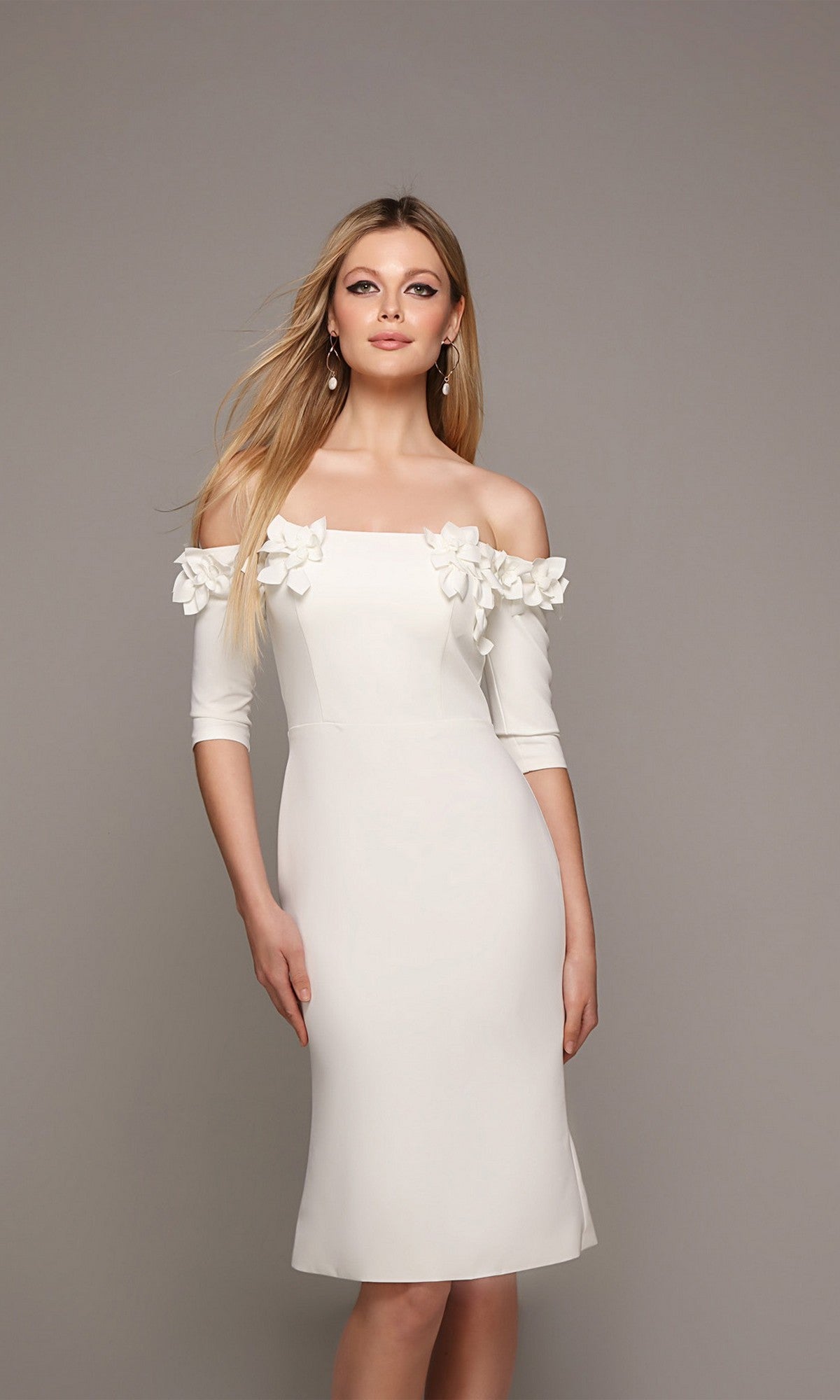 A woman wearing the Knee Length Semi-Formal Dress: Alyce 70003—an off-the-shoulder white dress featuring three-quarter sleeves and delicate 3D floral appliques—stands against a plain gray background.