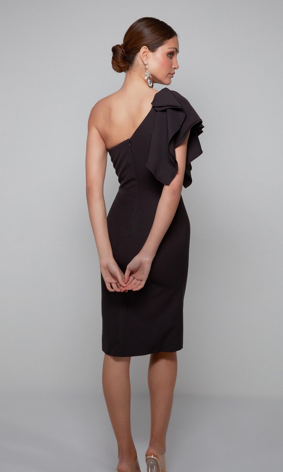 A woman faces away, wearing the Alyce 70005 knee length semi-formal dress with a one-shoulder ruffle detail and clear heels, set against a plain light gray background.
