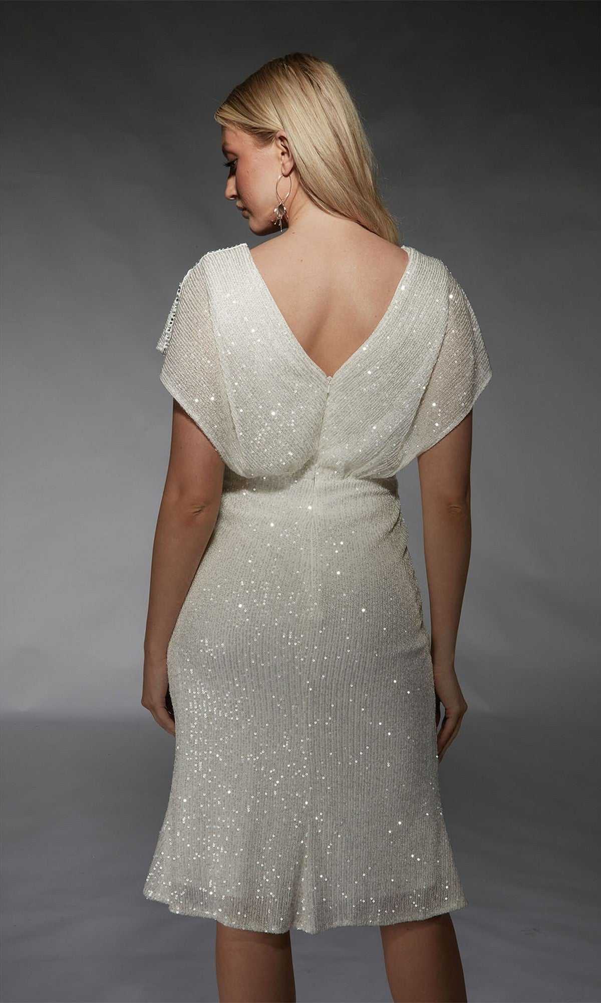 A woman with blonde hair faces away, wearing the Alyce 70063 Knee Length Semi-Formal Dress—a white, short-sleeved, sequin dress with a V-back. The background is plain gray.